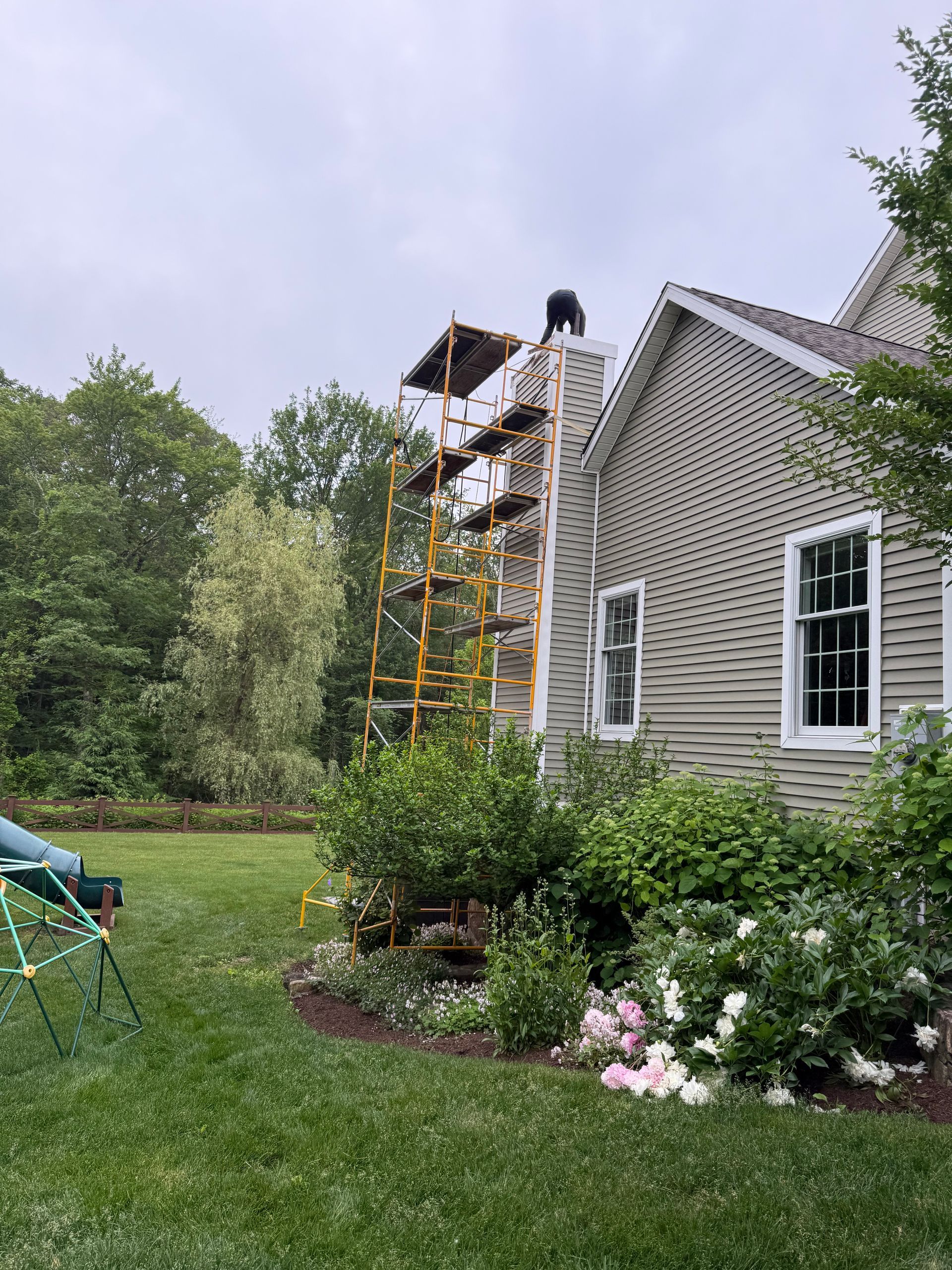 A person works on top of a yellow metal scaffold positioned against a brick chimney on the side of a house.