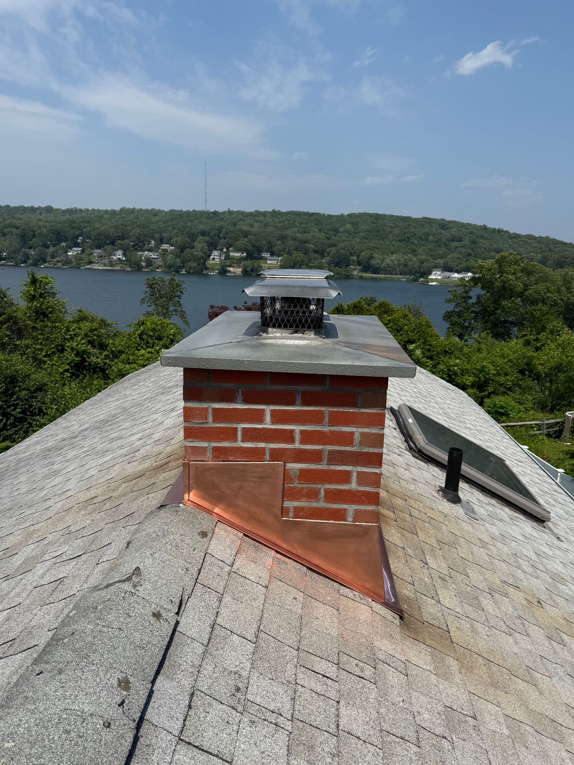 A brick chimney on a shingled roof with copper flashing, overlooking a lake surrounded by trees under a blue sky.