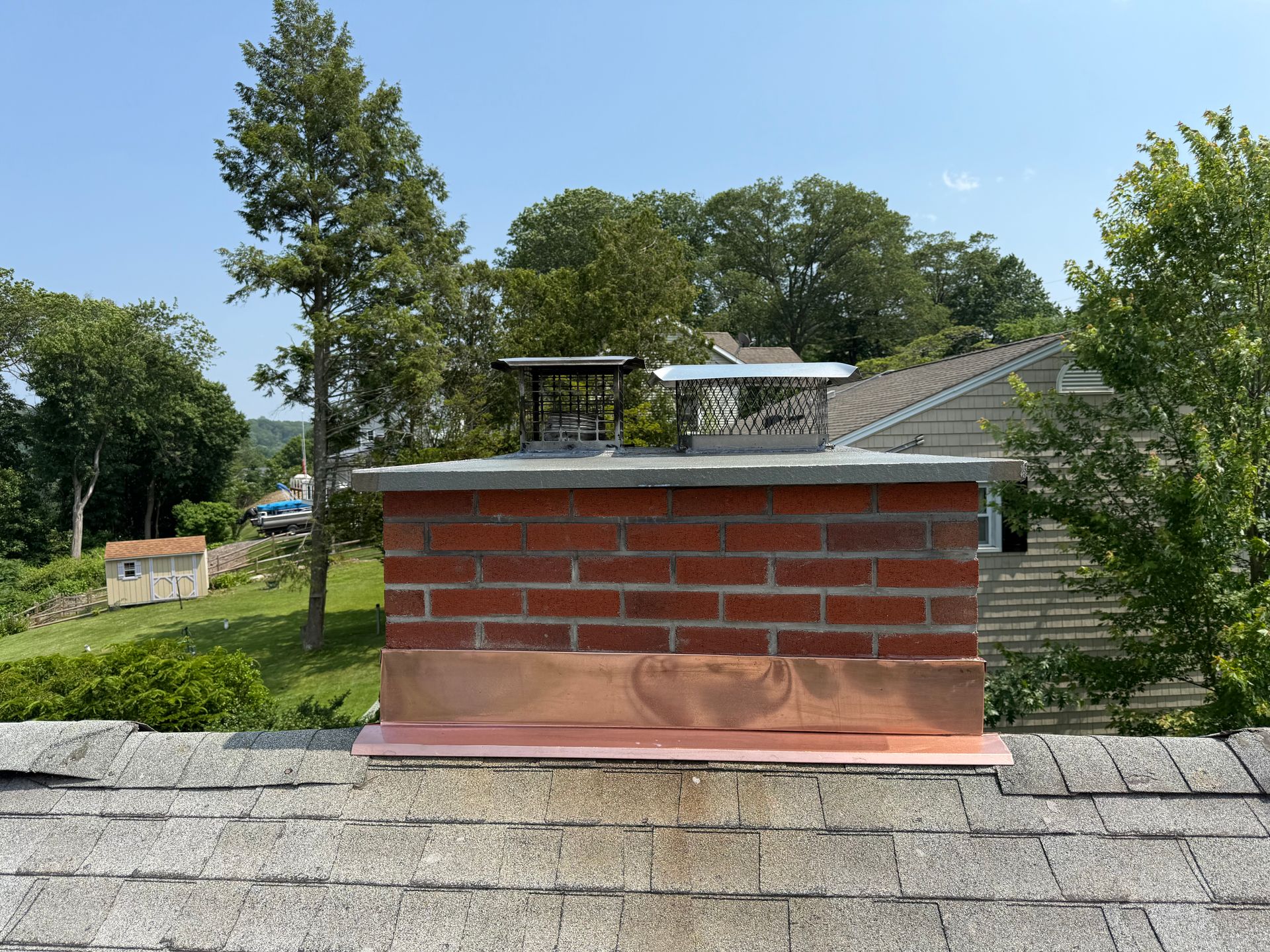 A brick chimney on a shingled roof with new copper flashing, topped with two metal chimney caps.