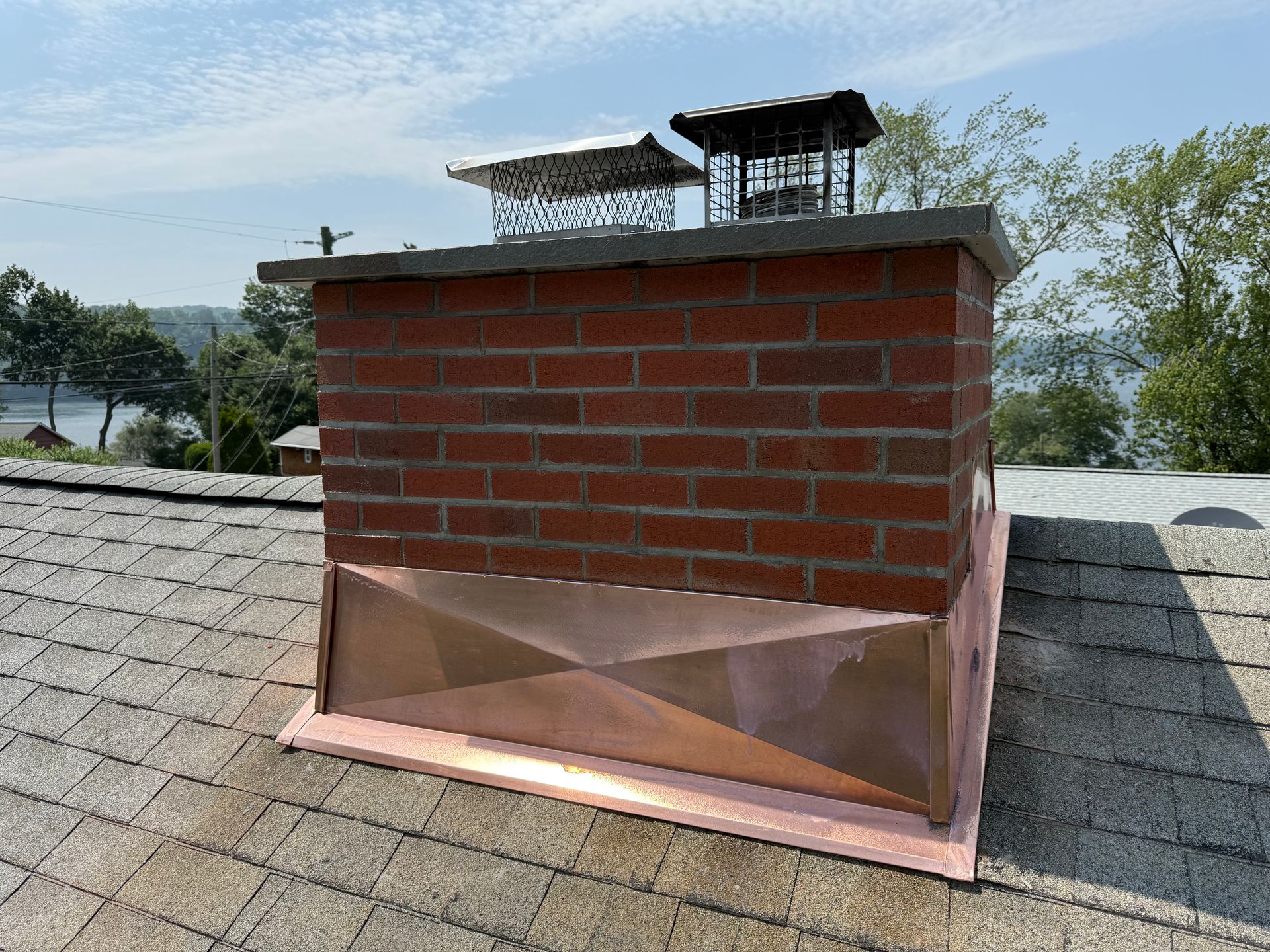 A brick chimney on a shingled roof features a new copper base flashing, with two metal chimney caps on top.