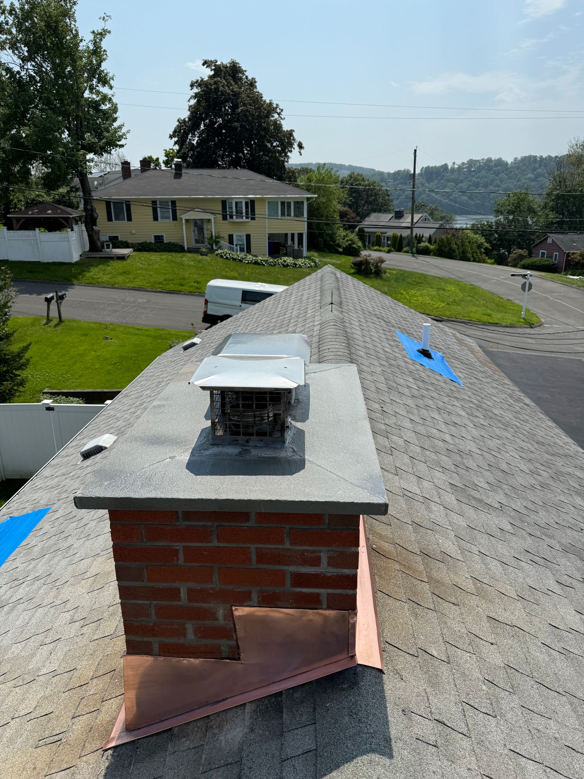A brick chimney on a shingled residential roof with copper flashing and a metal cap, viewed from above.