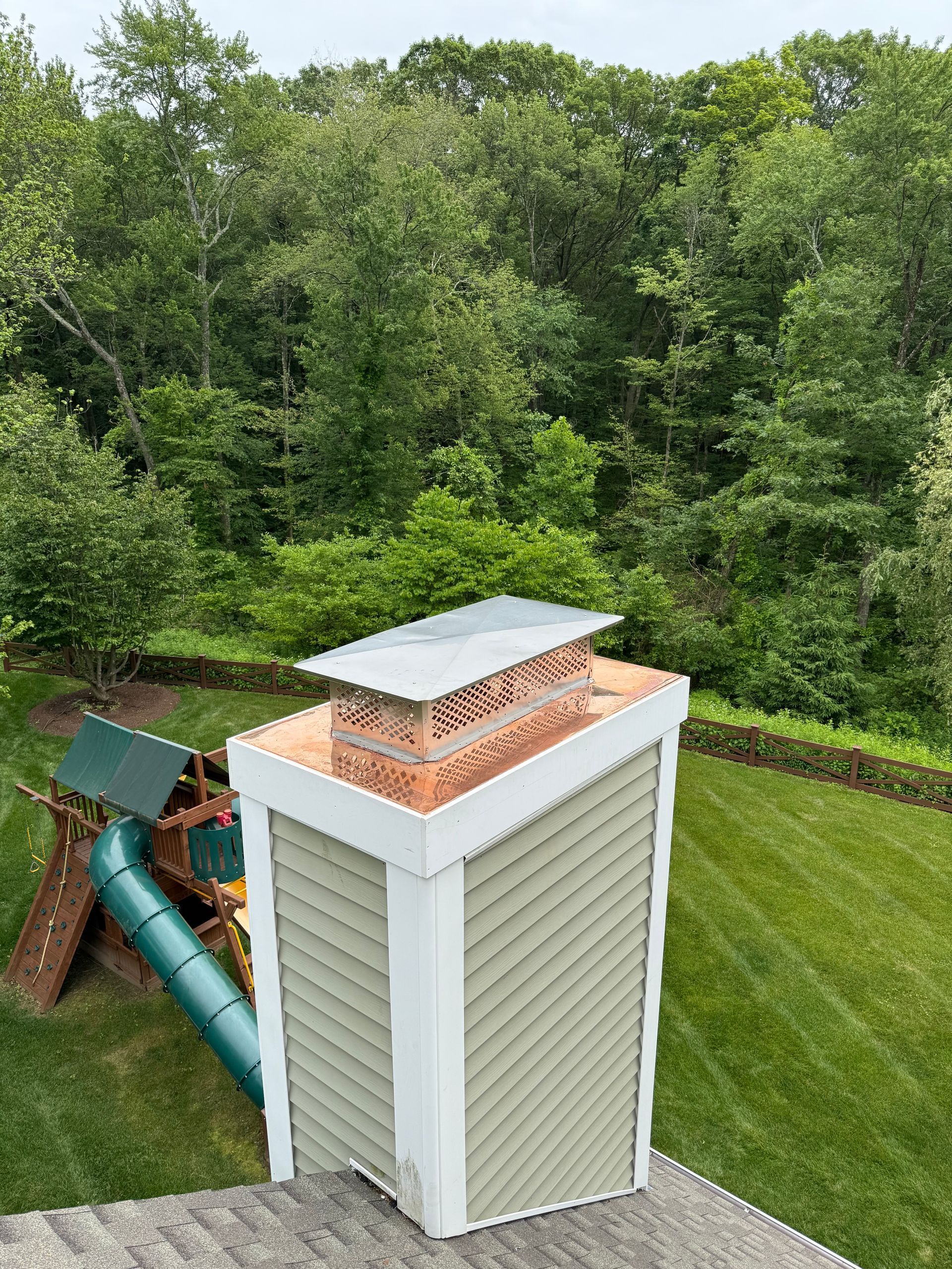 A chimney with green siding, white trim, and a metal cap on a roof, overlooking a green lawn and trees.