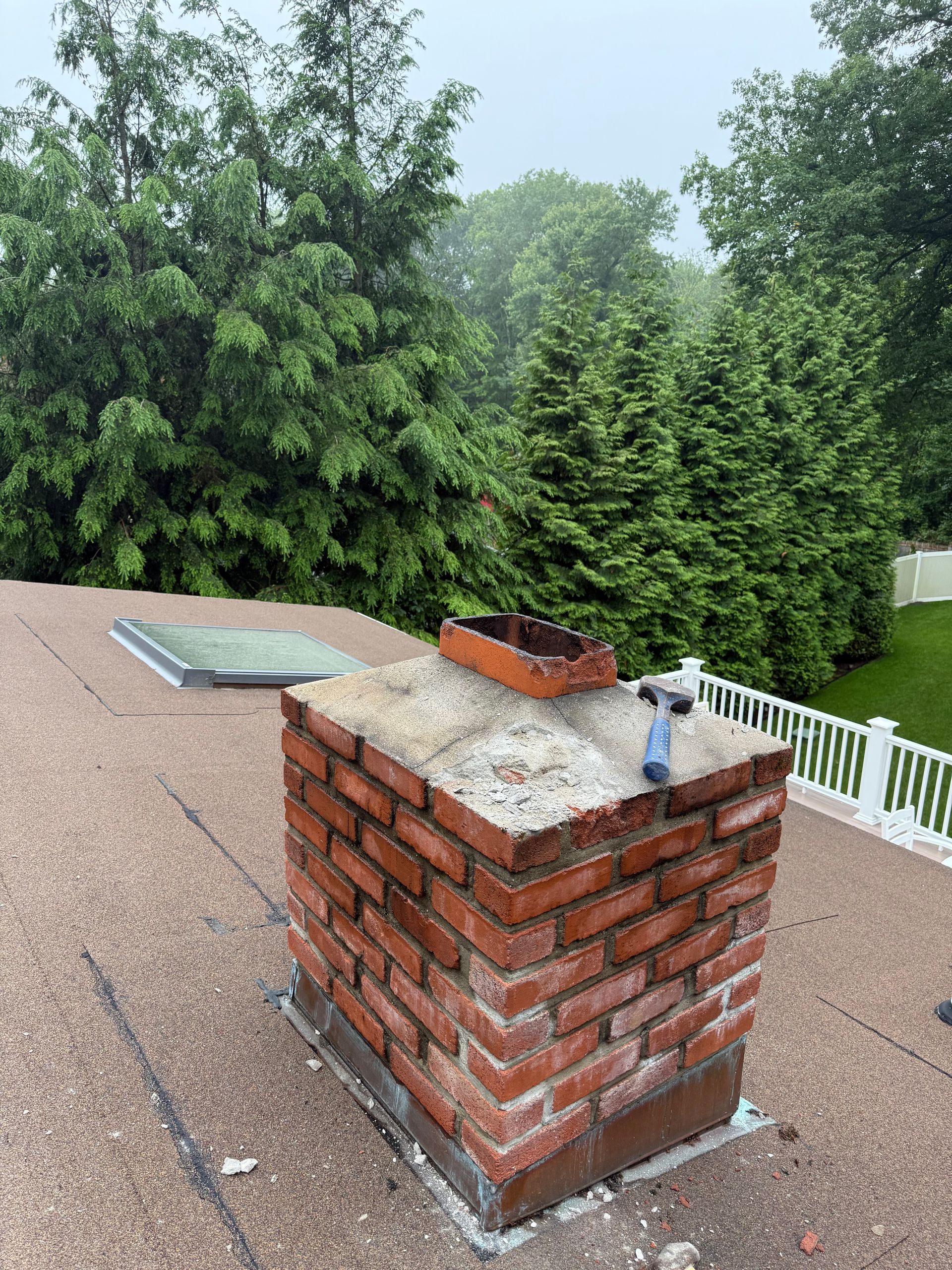 A brick chimney sits on a sloped shingled roof with a skylight nearby, featuring a blue hammer resting on its concrete cap.