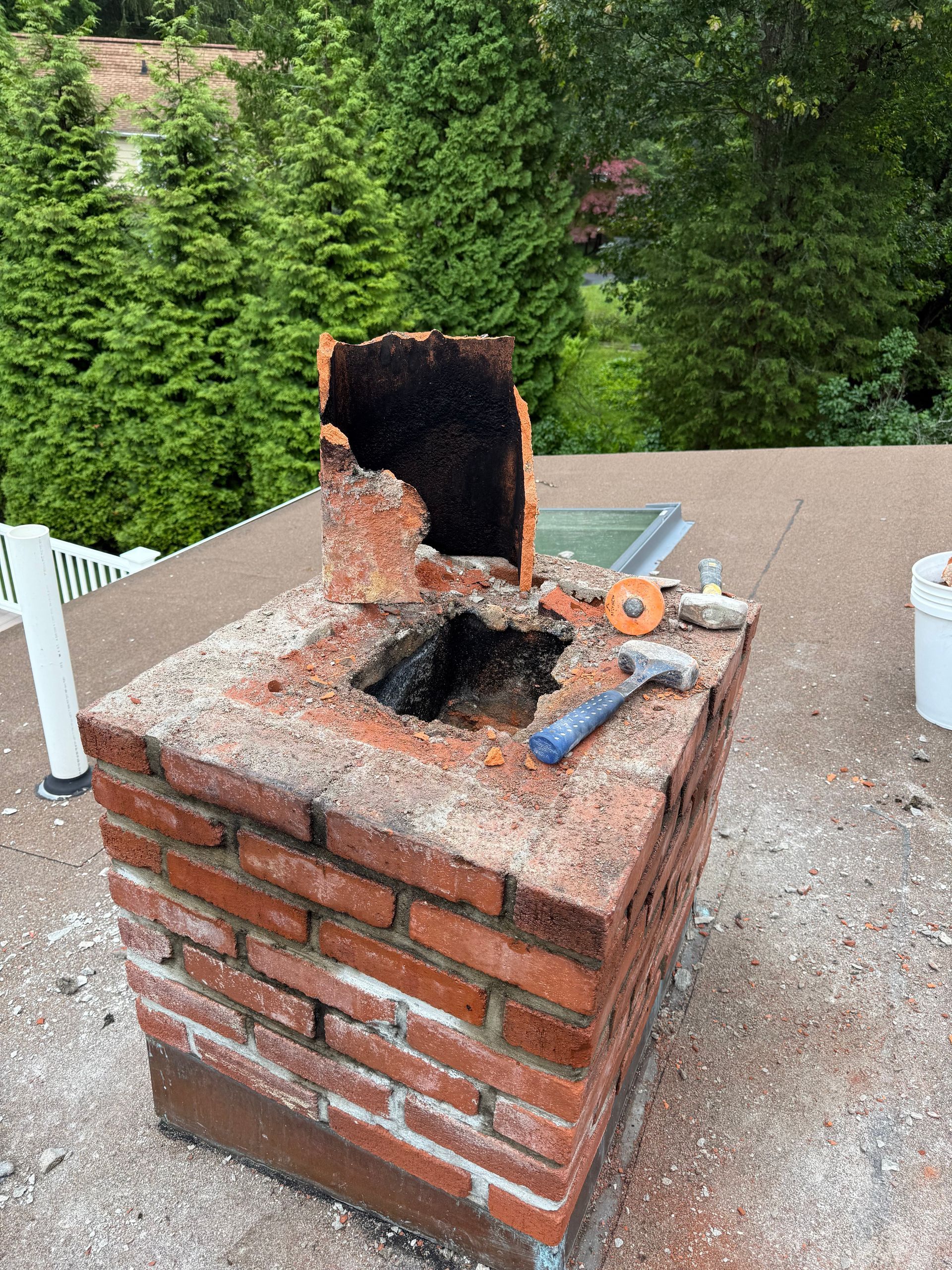 A brick chimney on a roof with a broken clay flue liner and a hammer lying on top.