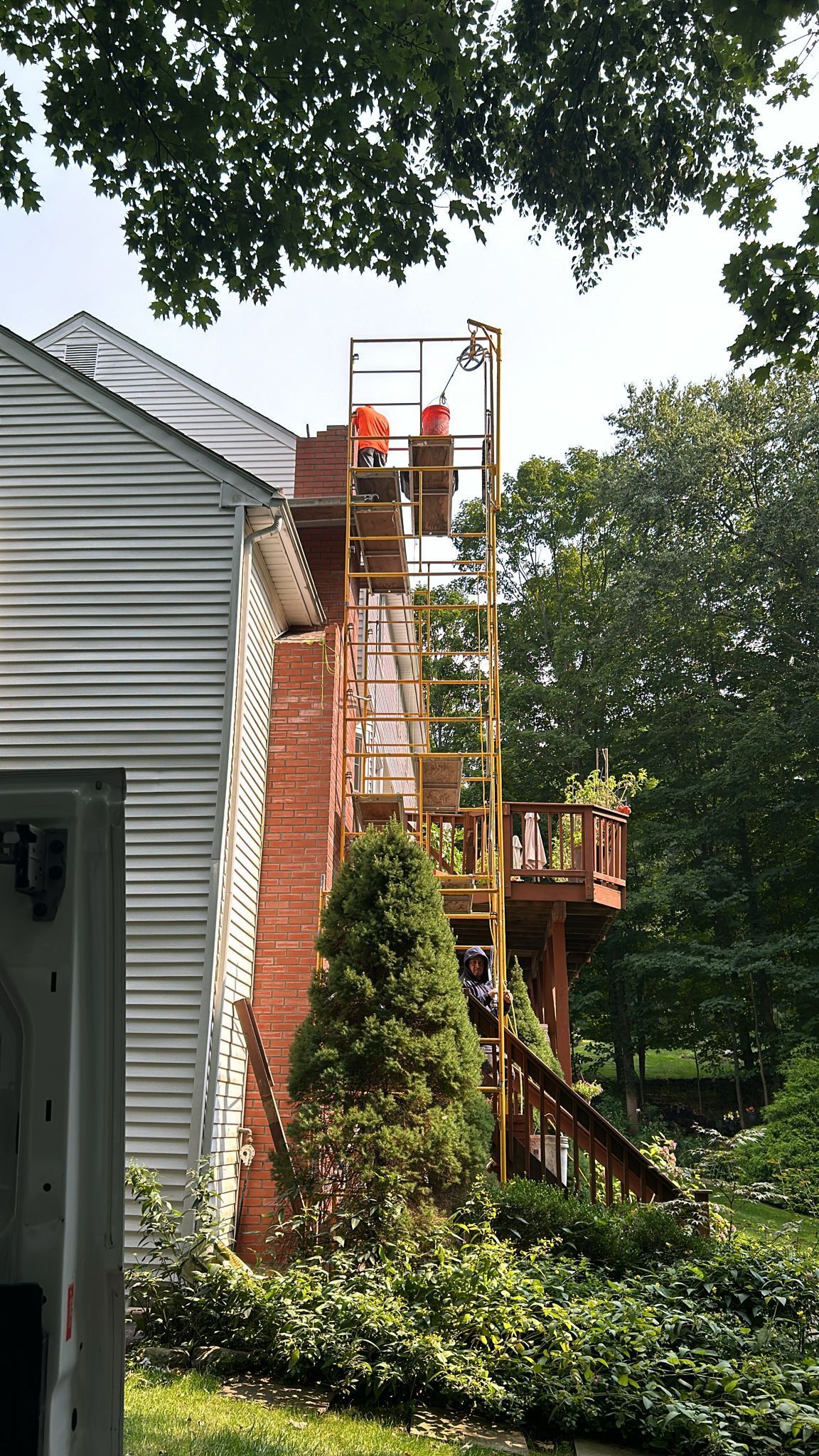 A worker in a high-visibility orange vest stands on a tall, narrow scaffold repairing a red brick chimney.