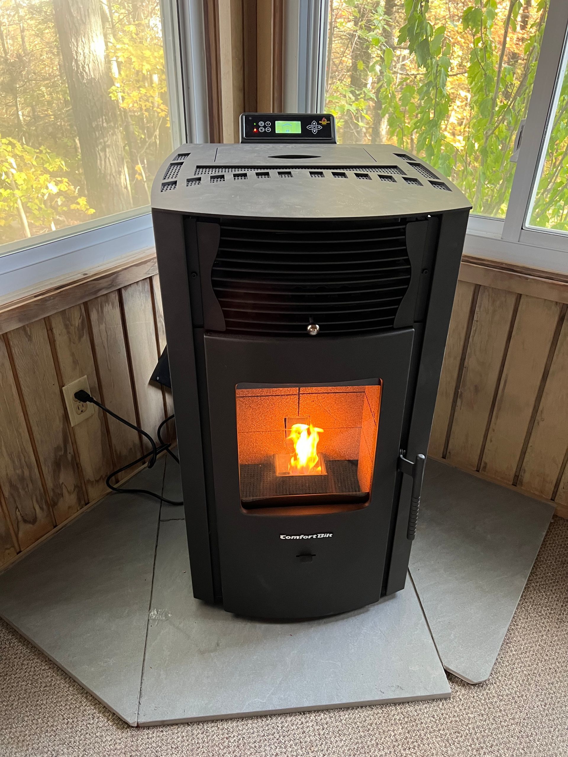 A black pellet stove with a glowing fire inside, placed on a hearth pad in a corner room with large windows.