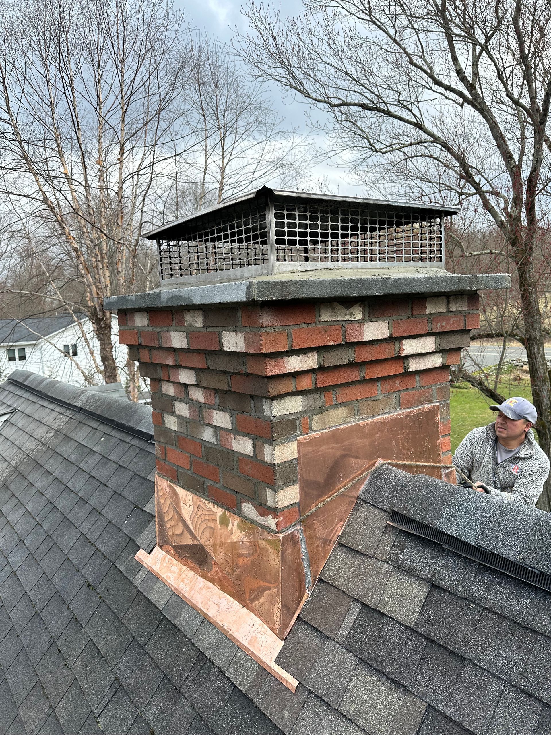 A worker repairing brick chimney flashing on a residential roof with a metal chimney cap.