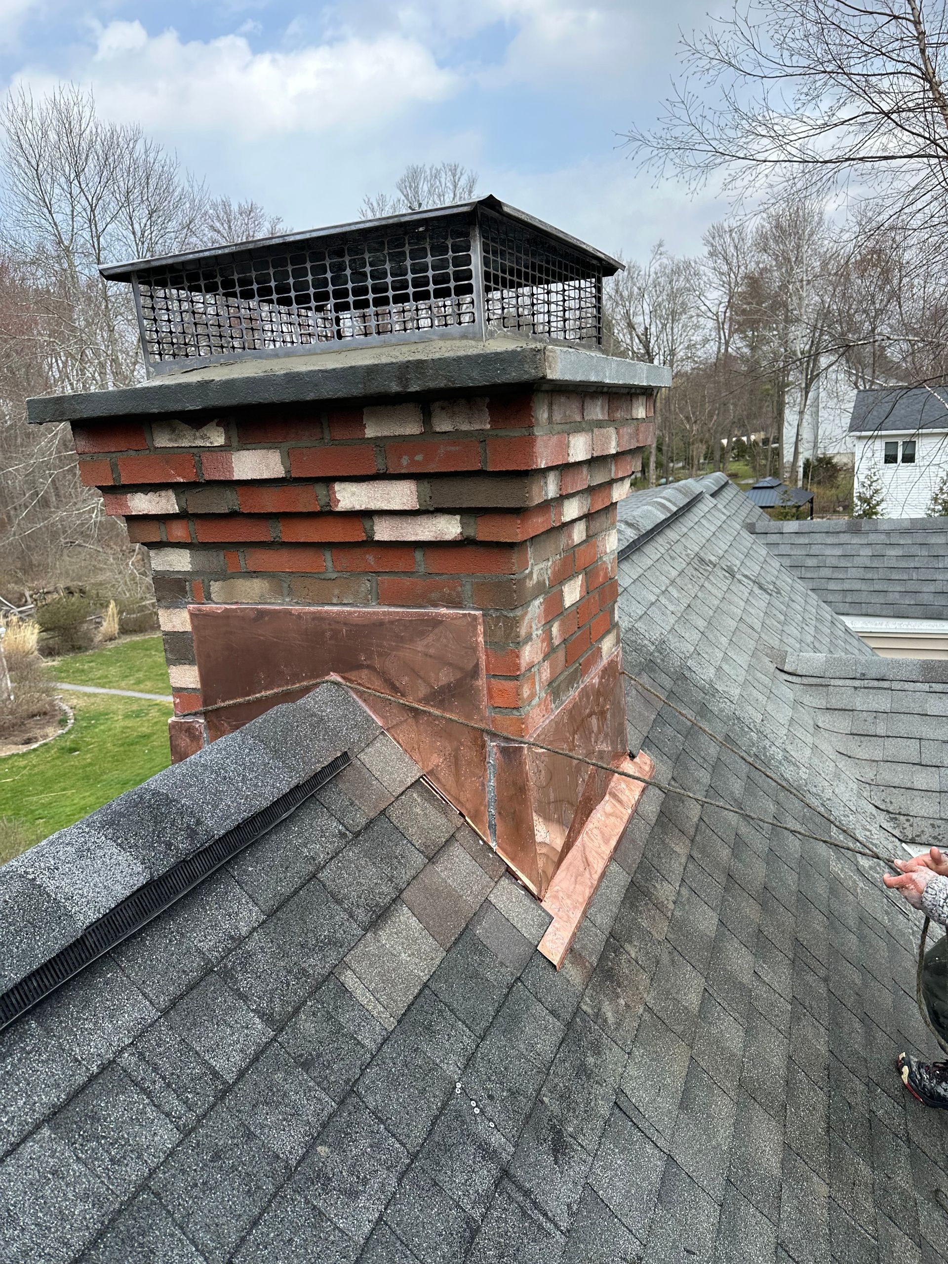 A brick chimney with a metal cap stands on a sloped, shingled roof, featuring copper flashing at the base.