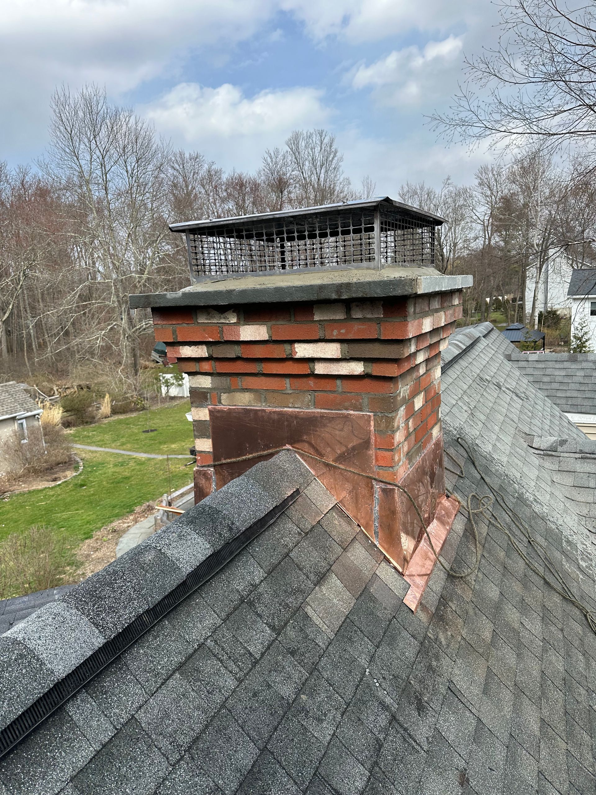 A red brick chimney topped with a metal cap, situated on a dark shingled roof under a partially cloudy sky.