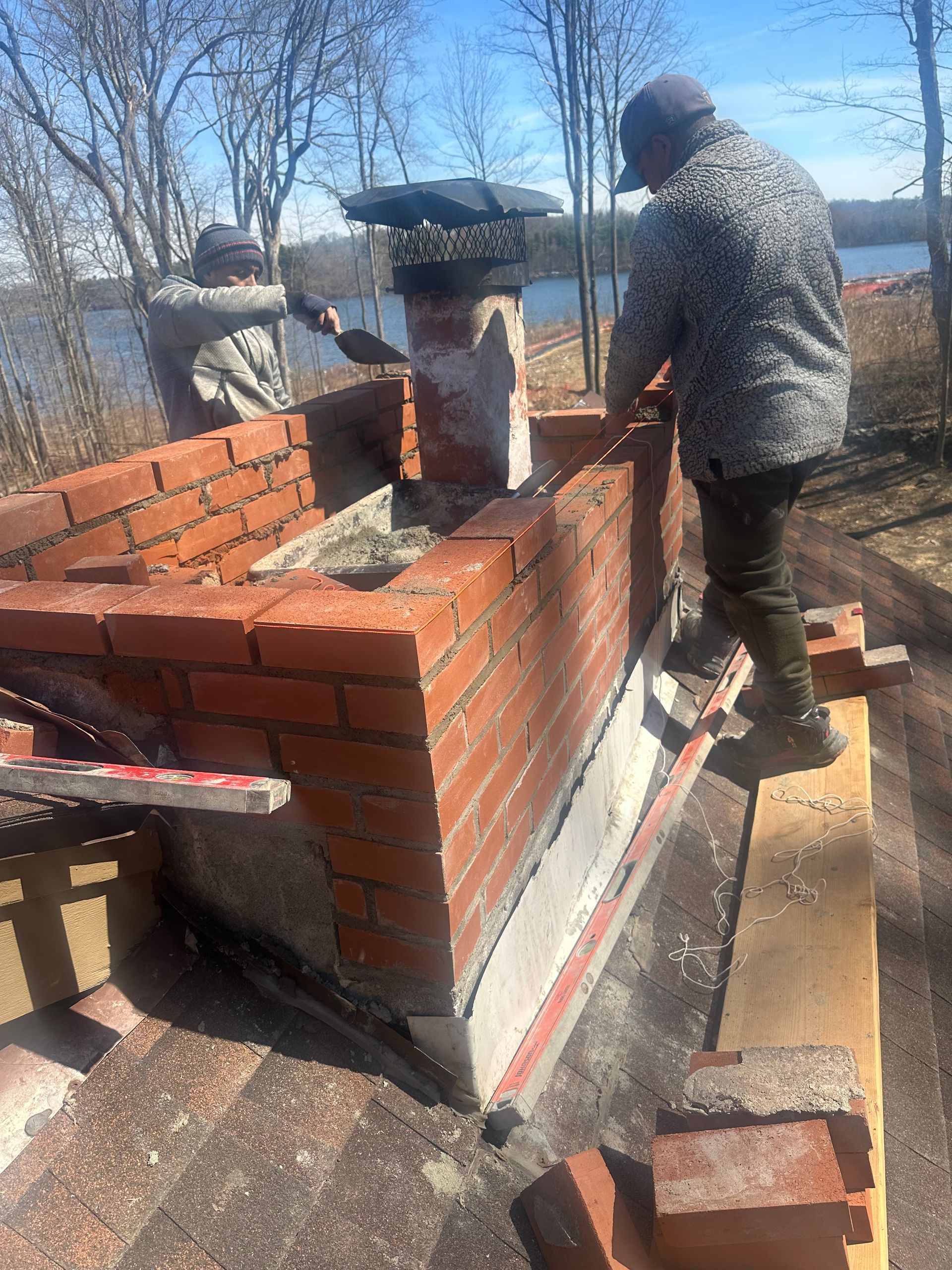 Two people repair a brick chimney on a sloped roof outdoors with a lake visible in the background.