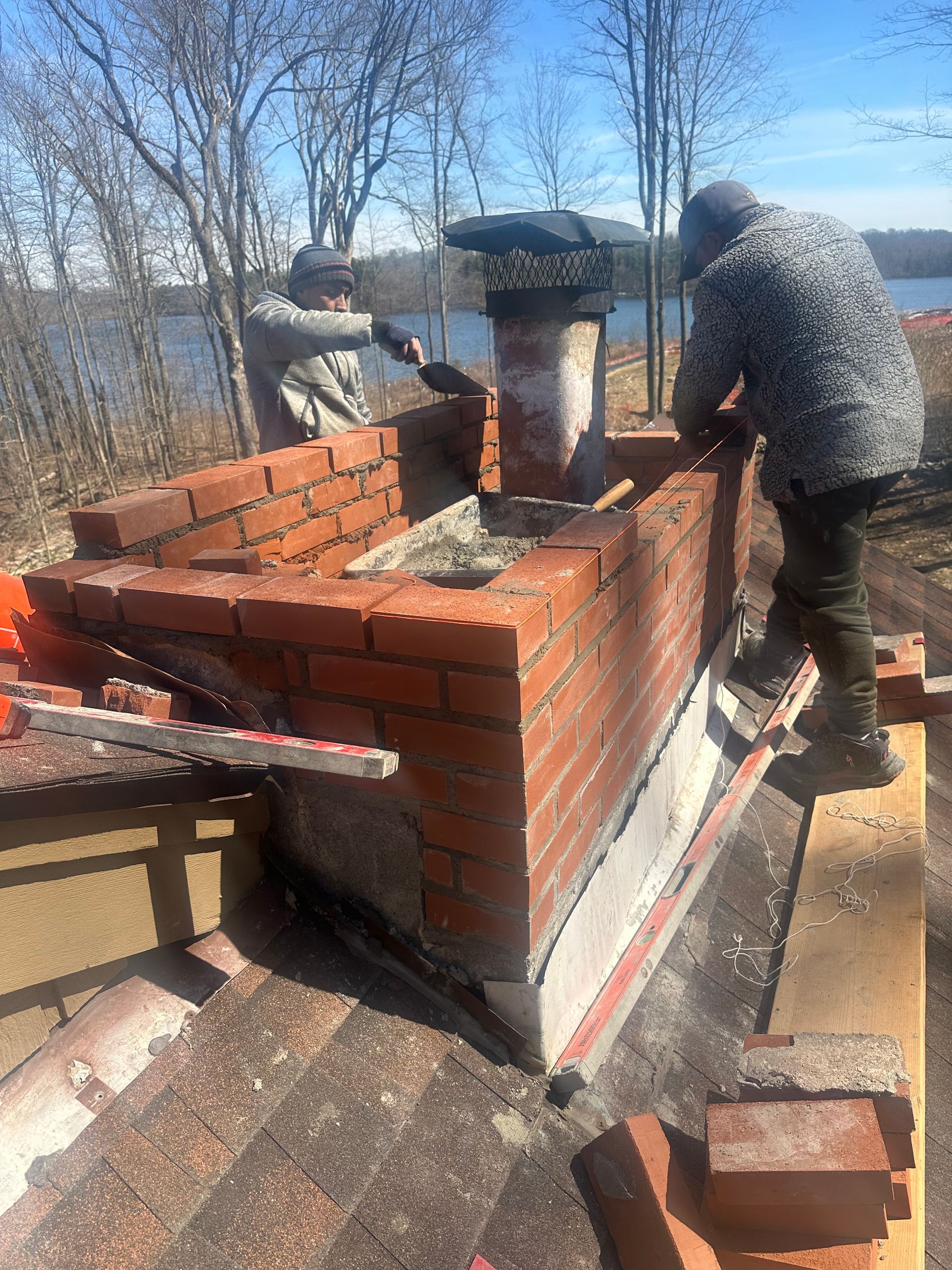 Two people work on a brick chimney repair on a roof, with trees and a lake in the background.