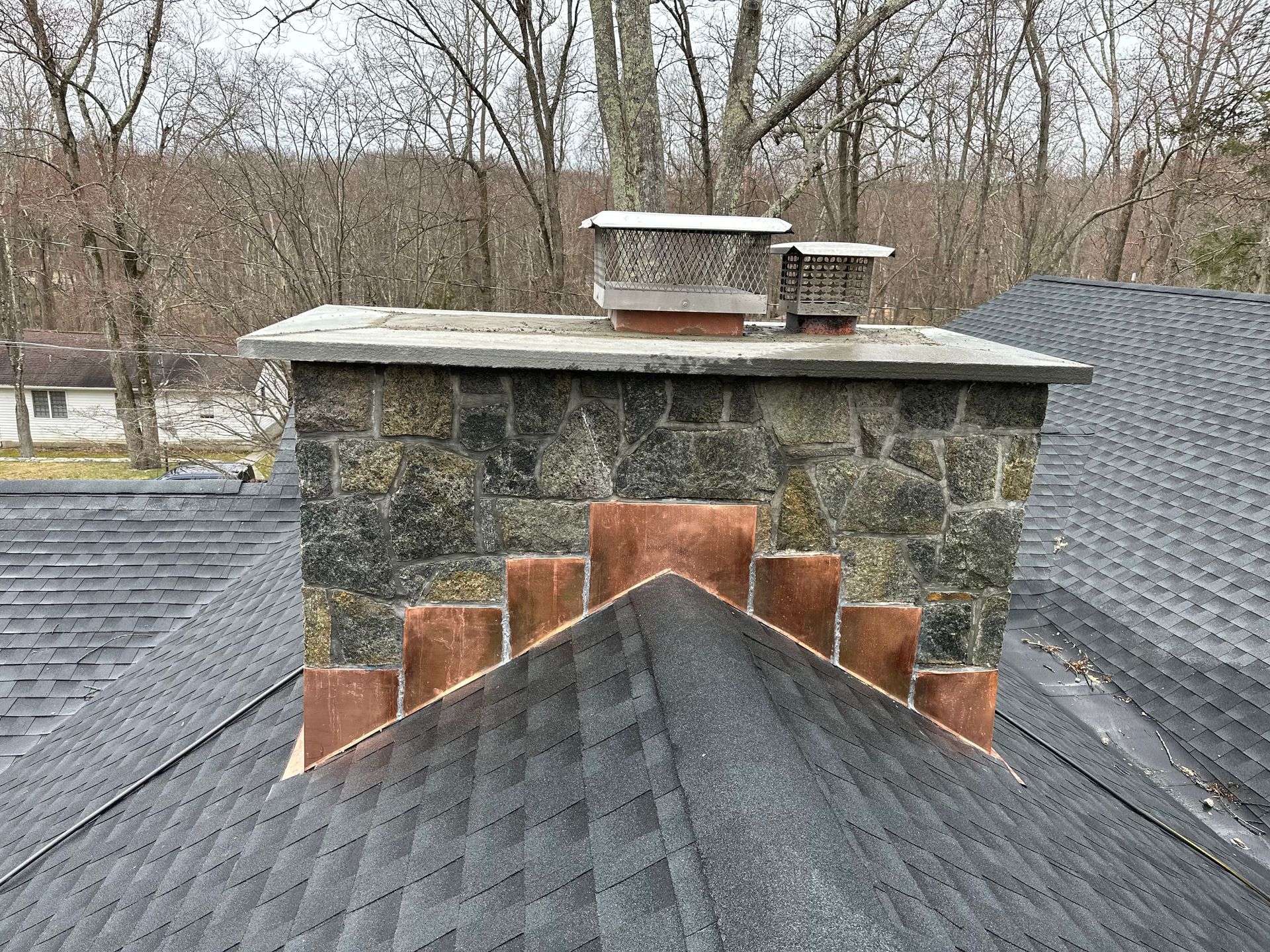 A stone chimney with a metal cap emerges from a shingled roof, showing a stepped base against a sloped roofline.