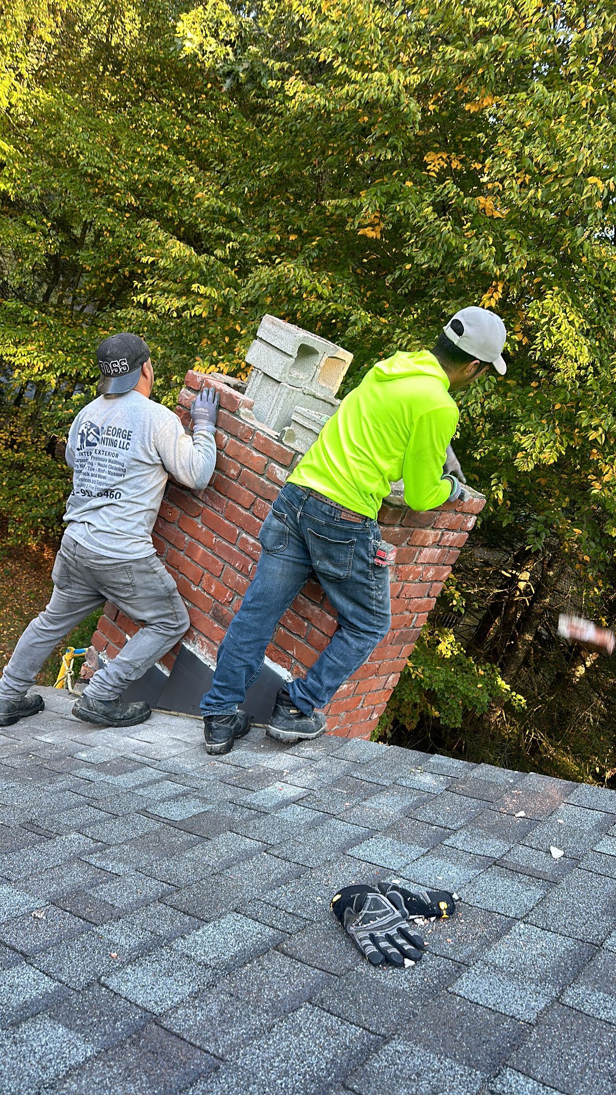 Two workers in work clothes and hats tilt a brick chimney structure atop a roof covered in shingles, outdoors.