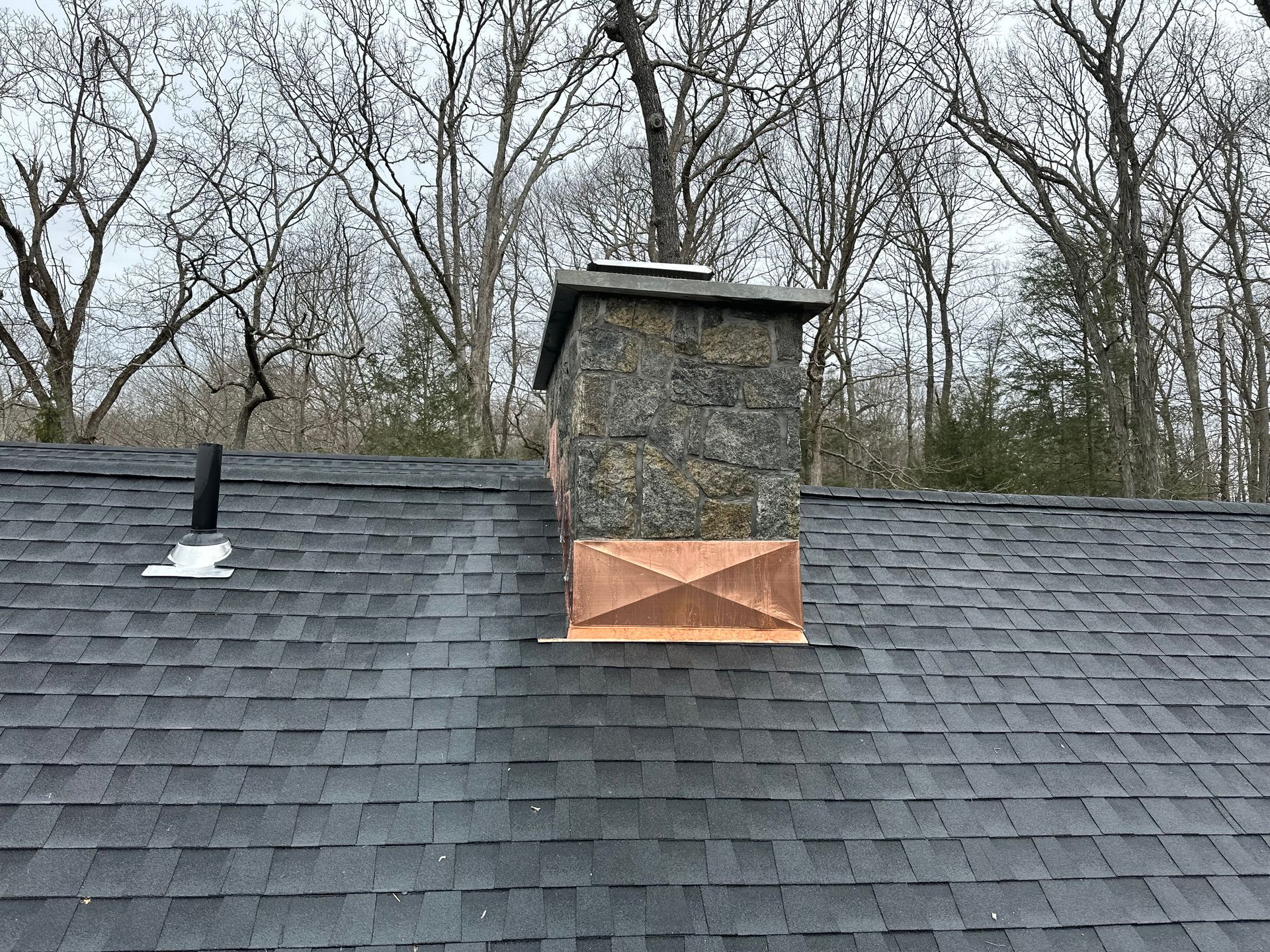 A dark stone chimney with a copper cricket on a shingled roof, set against a backdrop of bare winter trees.
