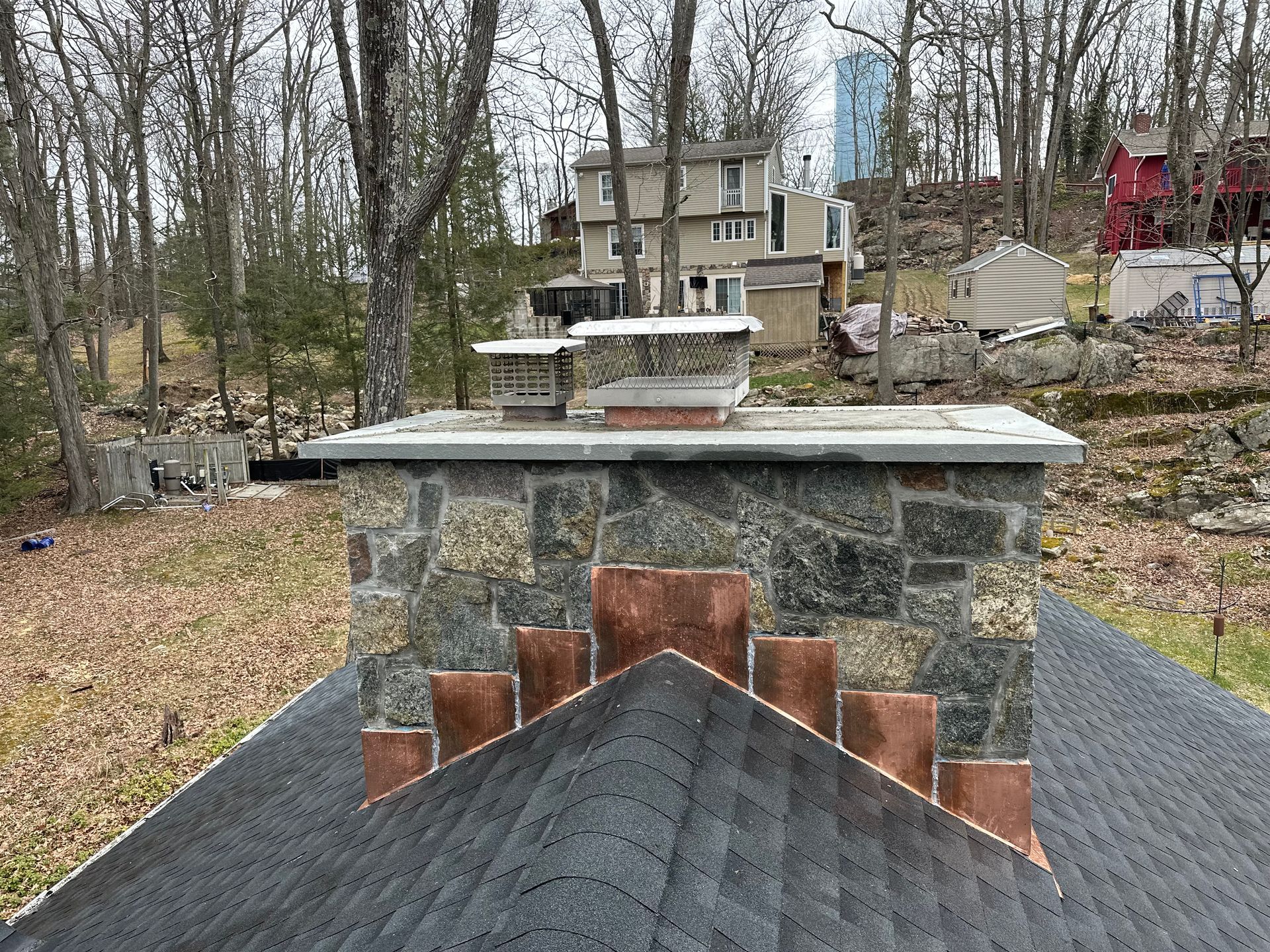 A stone chimney with copper flashing on a shingled roof, set against a backdrop of trees and a distant house.