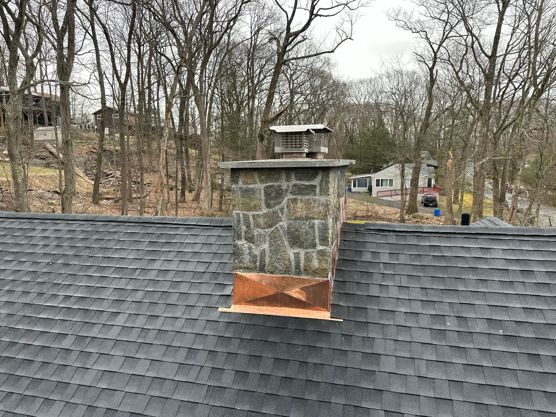 Stone chimney with copper flashing protruding from a dark gray shingled roof against a background of bare trees.