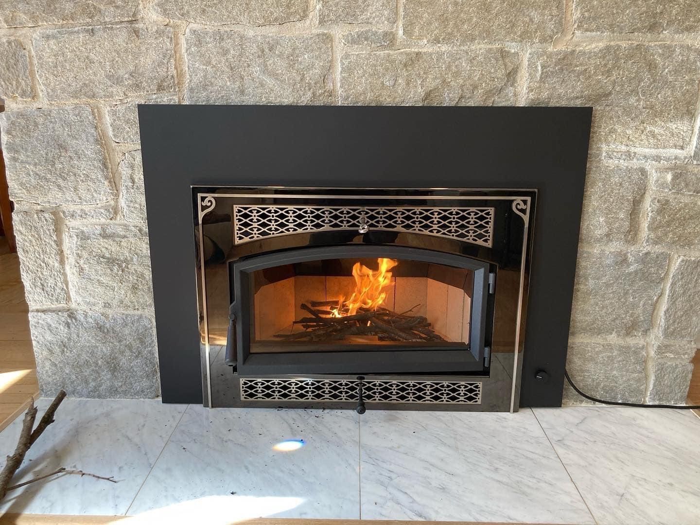 A lit wood-burning fireplace insert with a black frame installed in a textured gray stone fireplace.