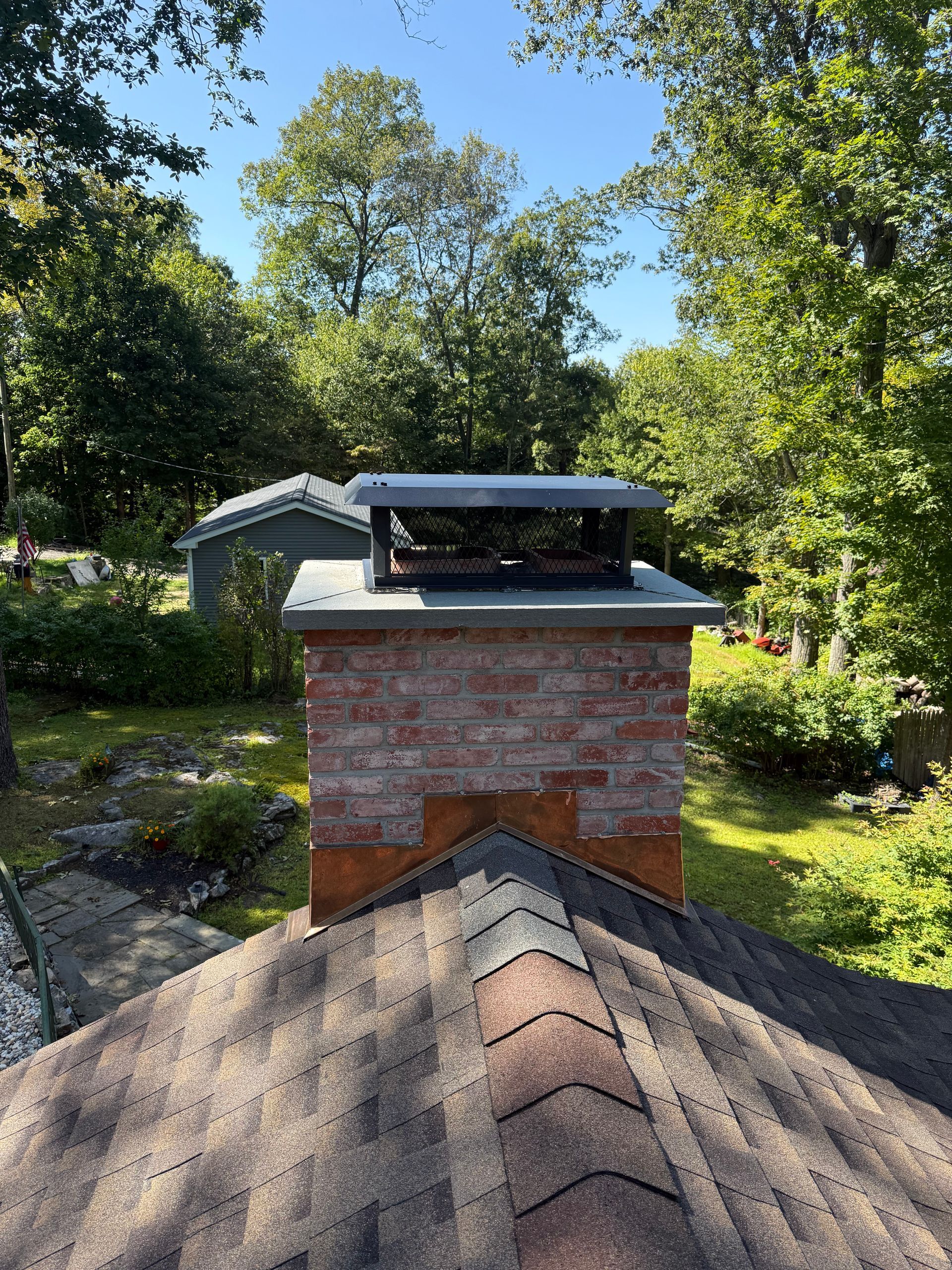 A brick chimney with a metal cap and screen sits on a shingled roof, surrounded by green trees on a sunny day.