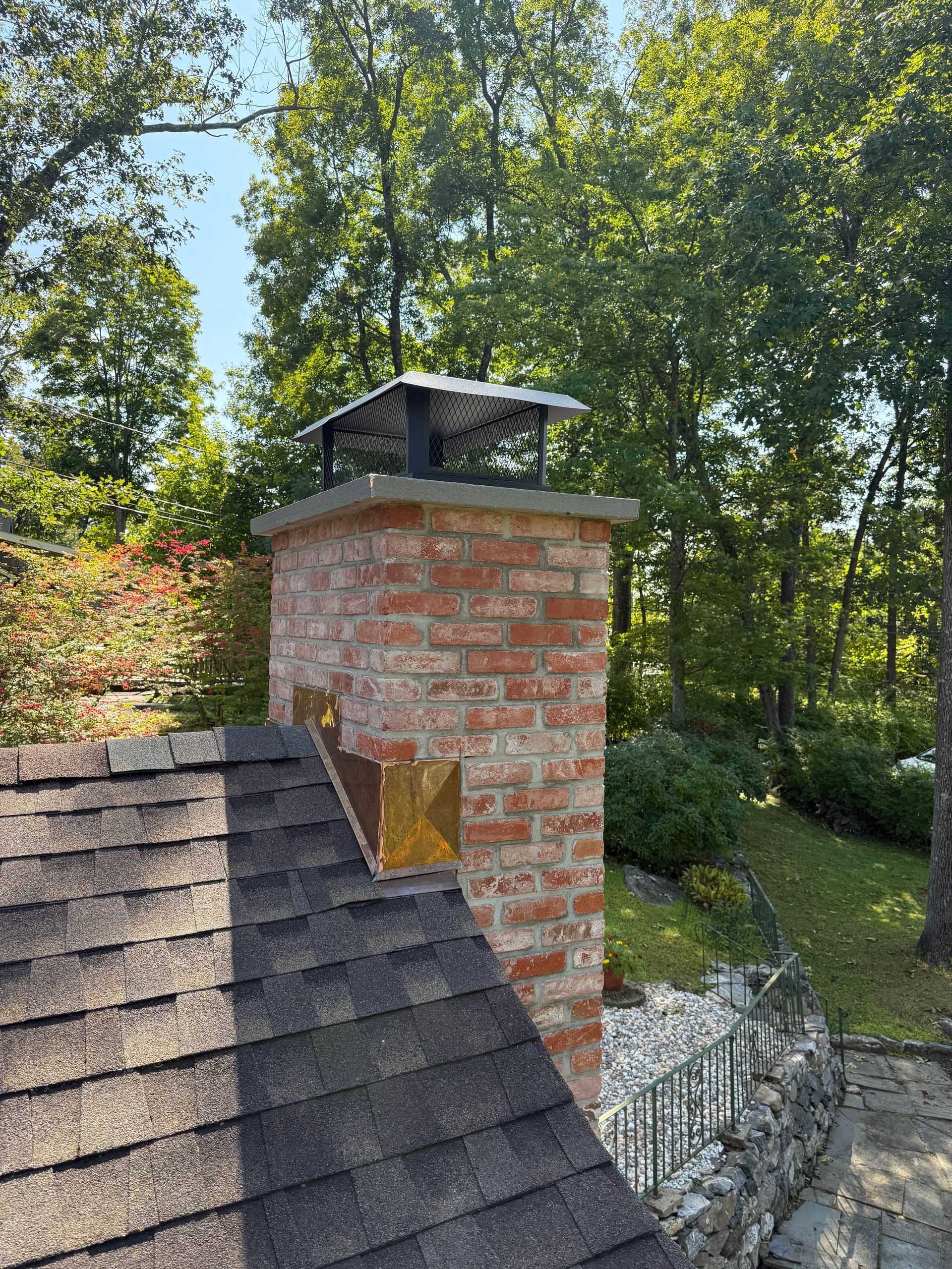 A brick chimney with a metal cap on a shingled roof, surrounded by trees and a stone wall.