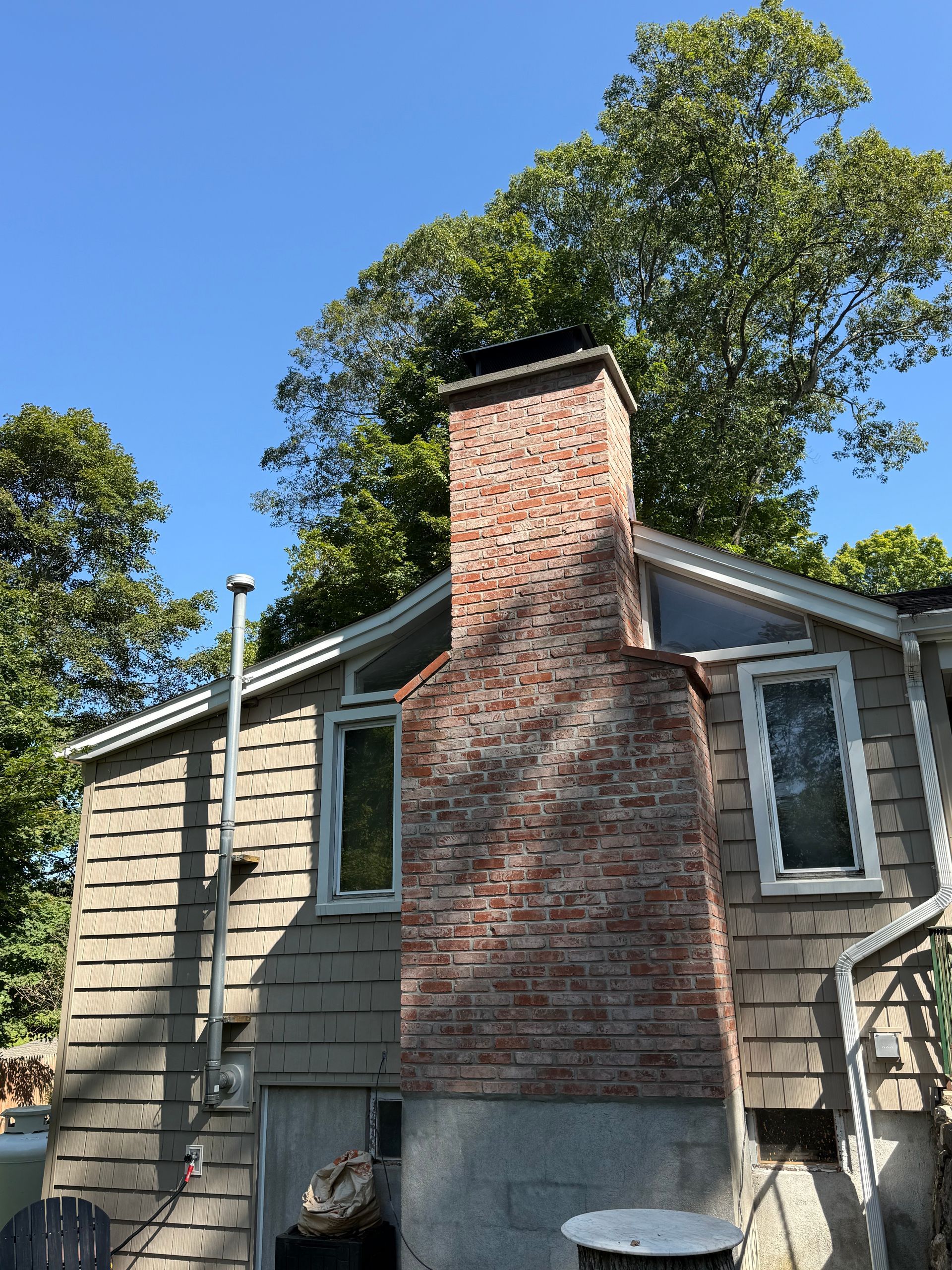 A large, weathered brick chimney stands against the side of a grey shingled house under a clear blue sky and leafy trees.