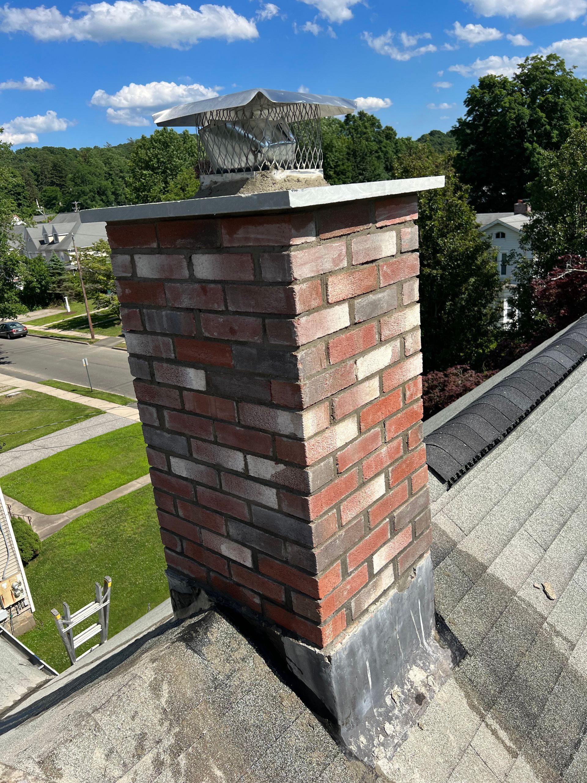 A brick chimney stands on a pitched roof under a blue sky with white clouds, topped with a metallic chimney cap.