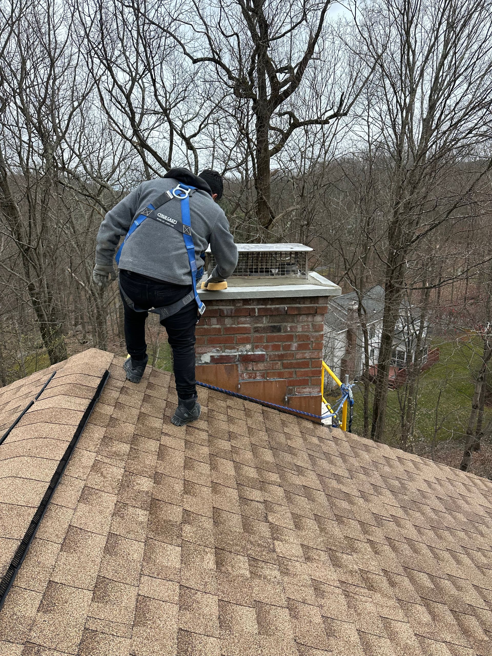 A roofer wearing a safety harness works on a shingled roof next to a brick chimney in a wooded area.