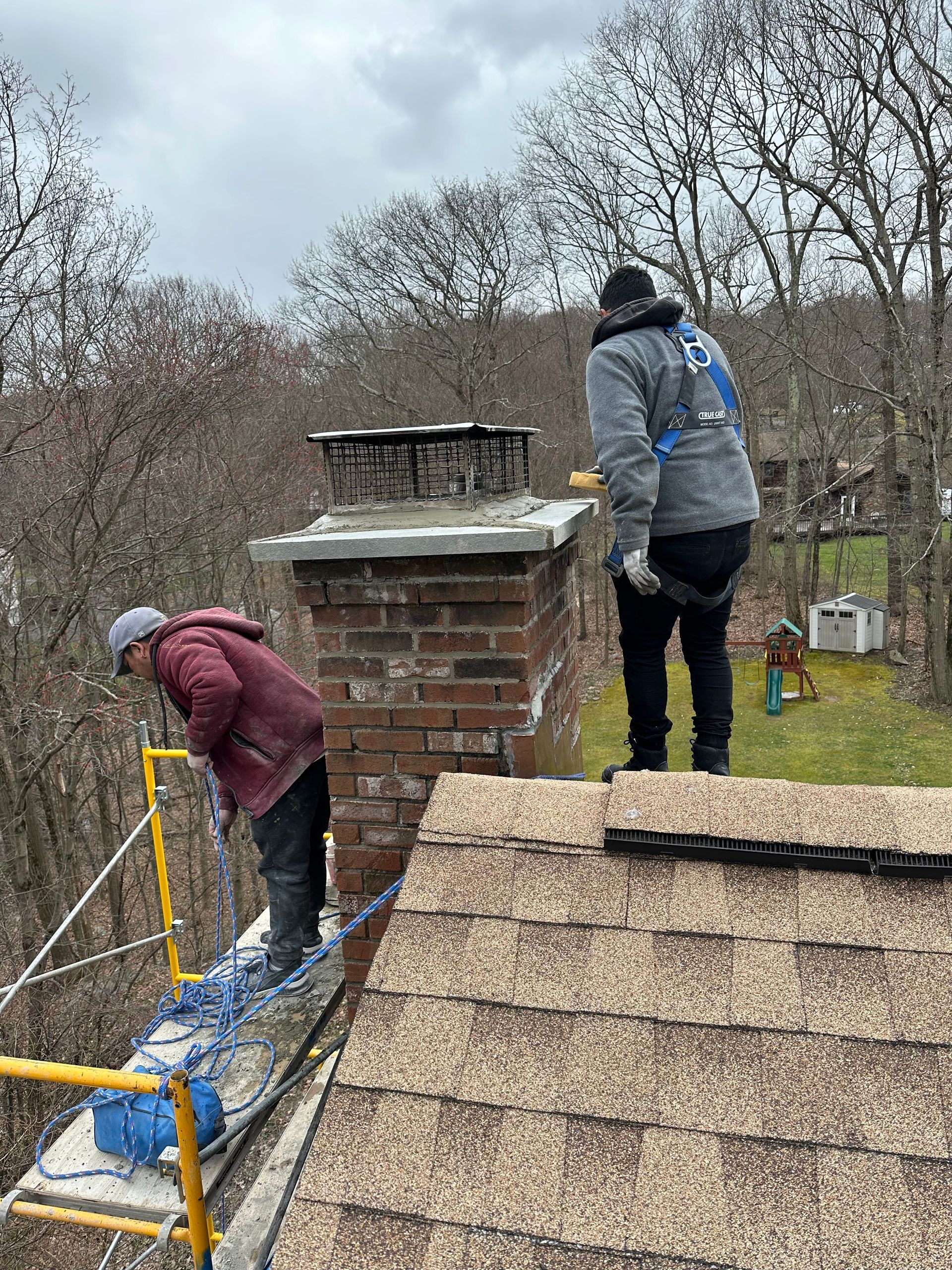 Two workers in safety harnesses perform masonry repairs on a brick chimney on a shingled roof.