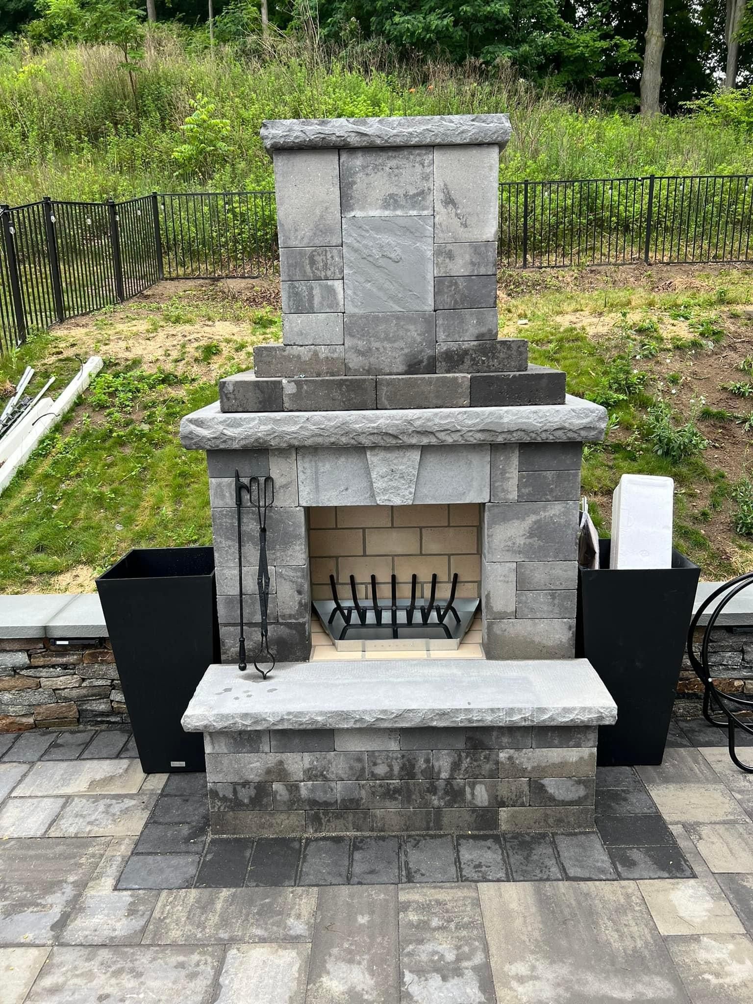 An outdoor stone fireplace with a fire grate and granite hearth, situated on a stone patio with black planters on sides.