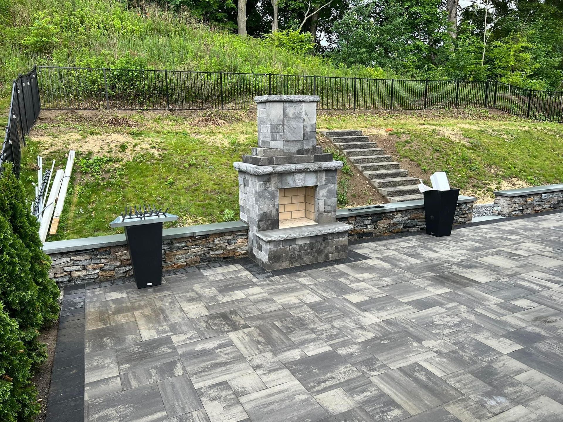 A stone outdoor fireplace sits on a gray paver patio next to black planters and stone steps leading up a grassy hill.