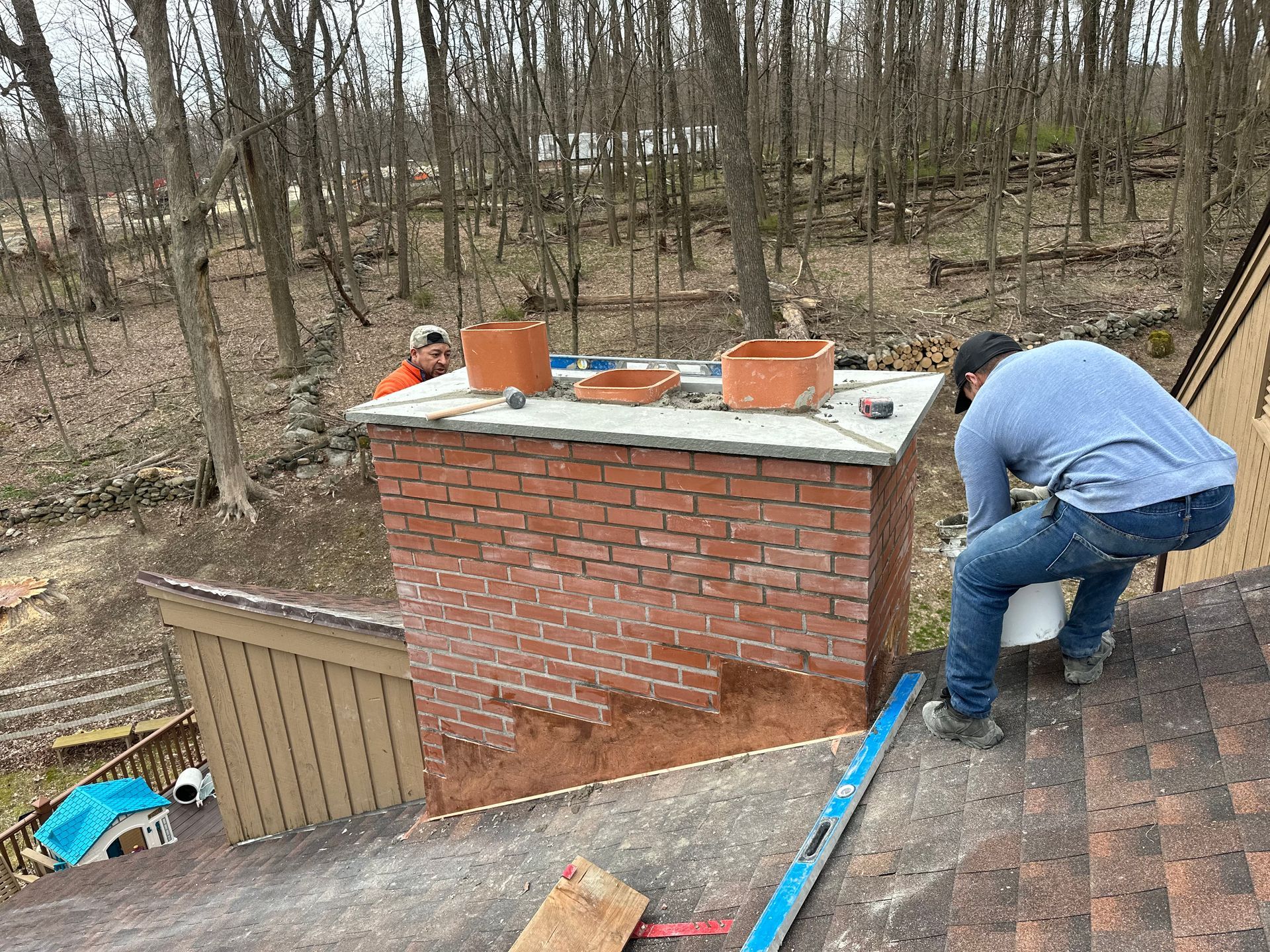 Two workers on a residential roof repair a brick chimney surrounded by a wooded area.