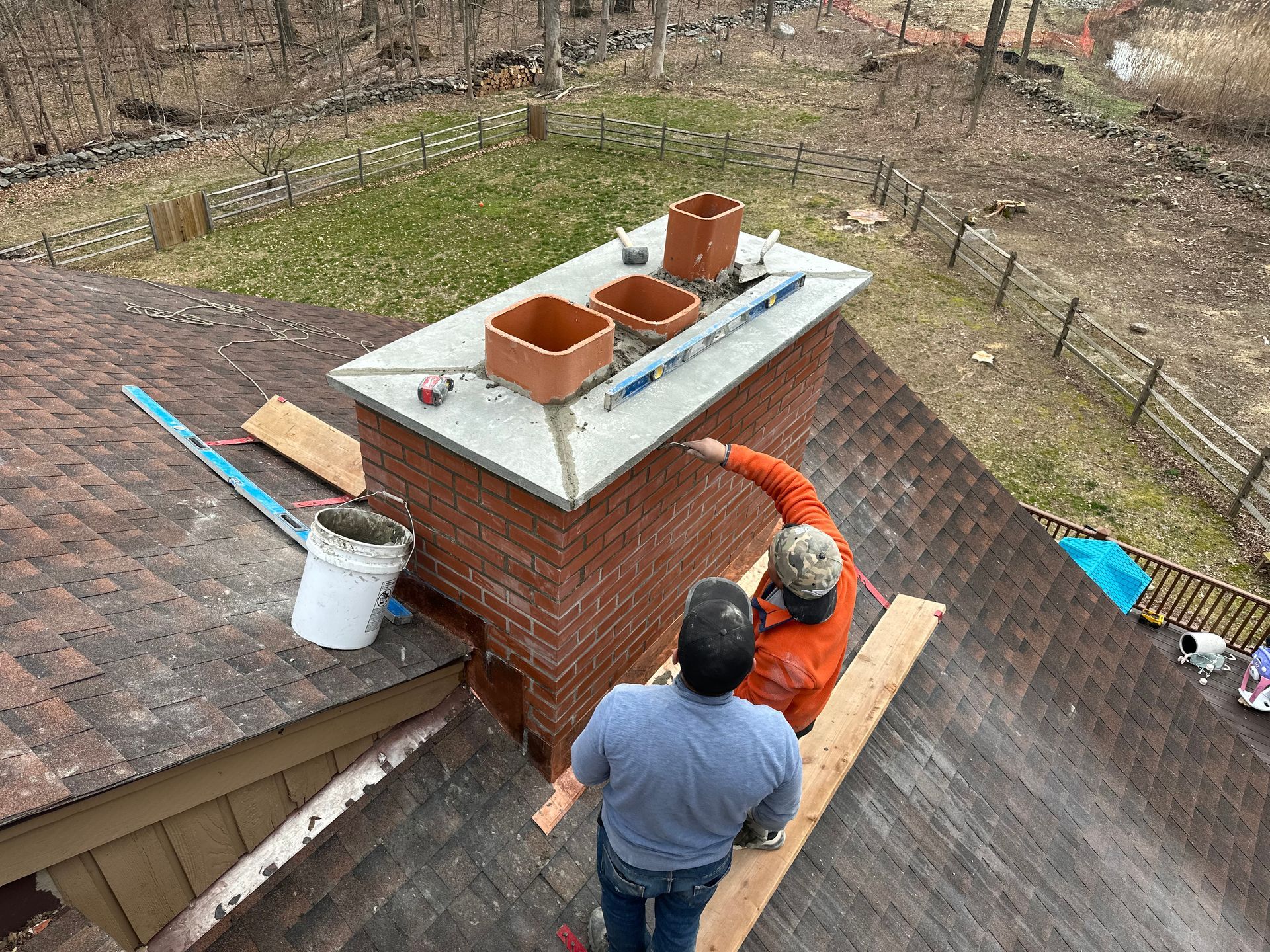 Two workers repairing a brick chimney on a shingled residential roof with a surrounding wooded yard.