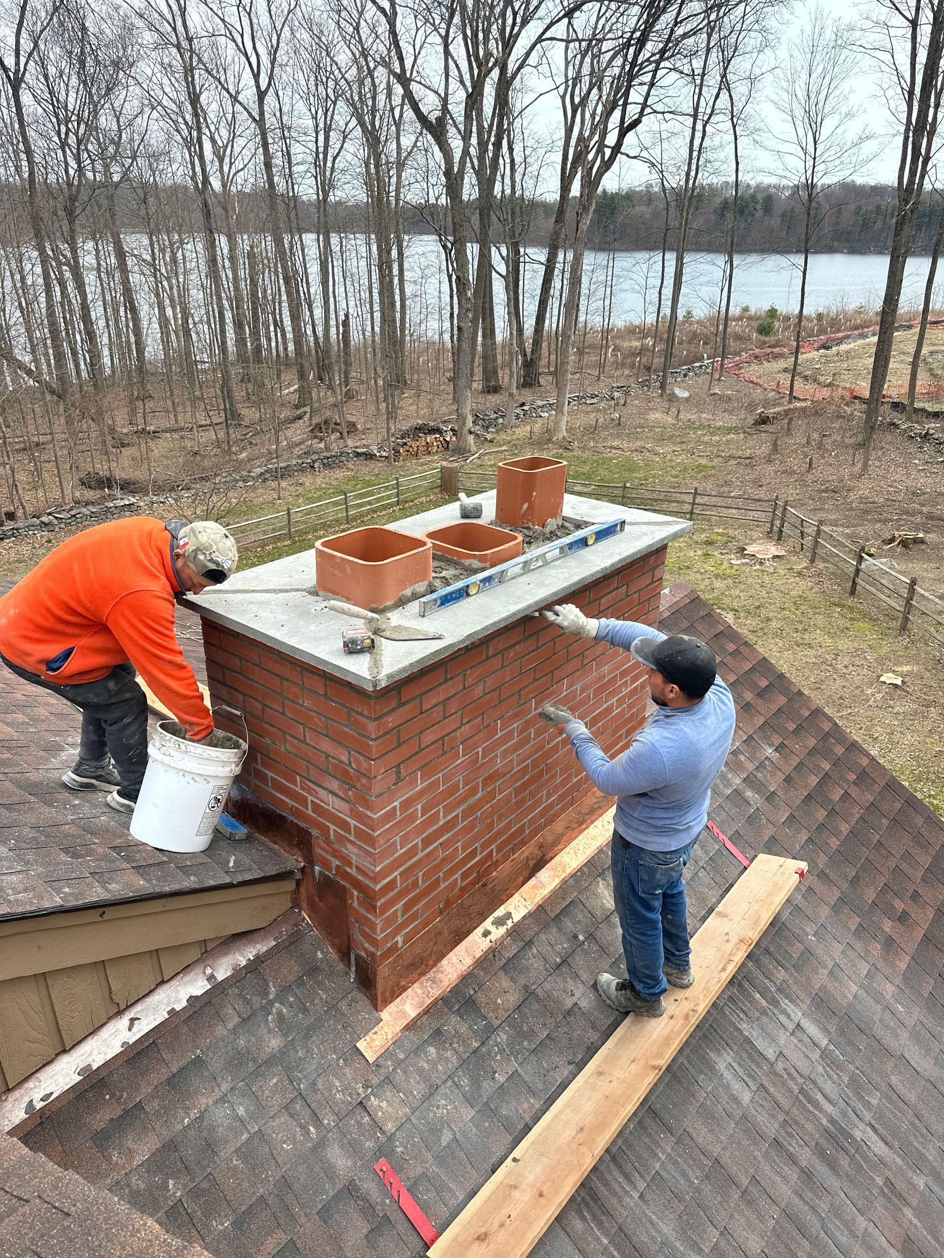 Two workers on a residential roof install a new concrete chimney cap on a brick chimney overlooking a lake.