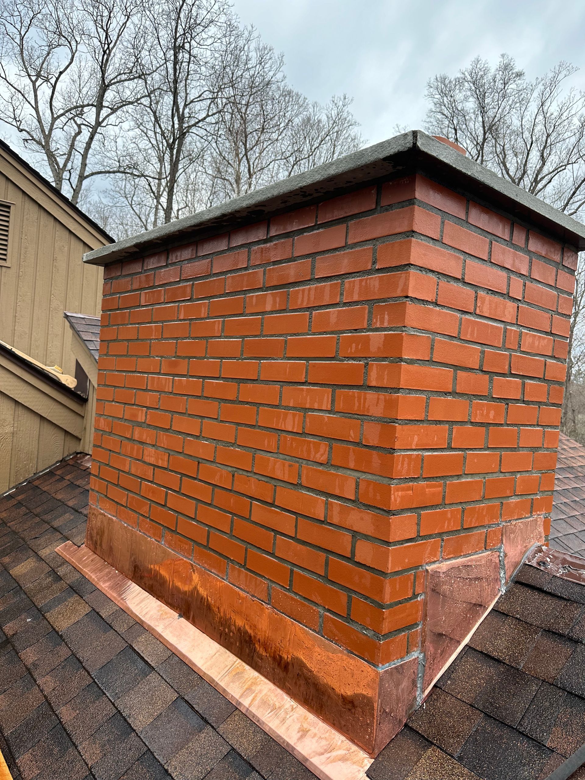 A brick chimney on a shingled roof with new copper flashing installed at the base.
