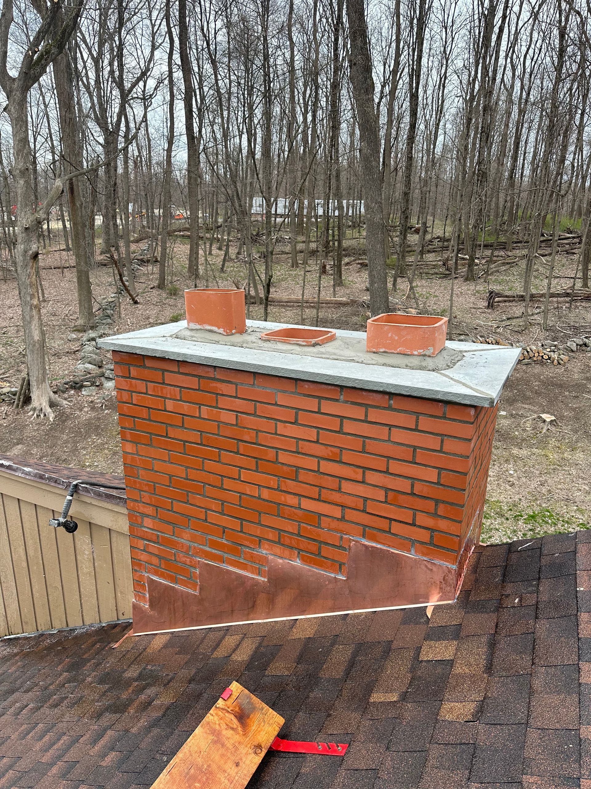 A red brick chimney on a shingled roof with copper flashing and three ceramic flue tiles, set against a wooded background.