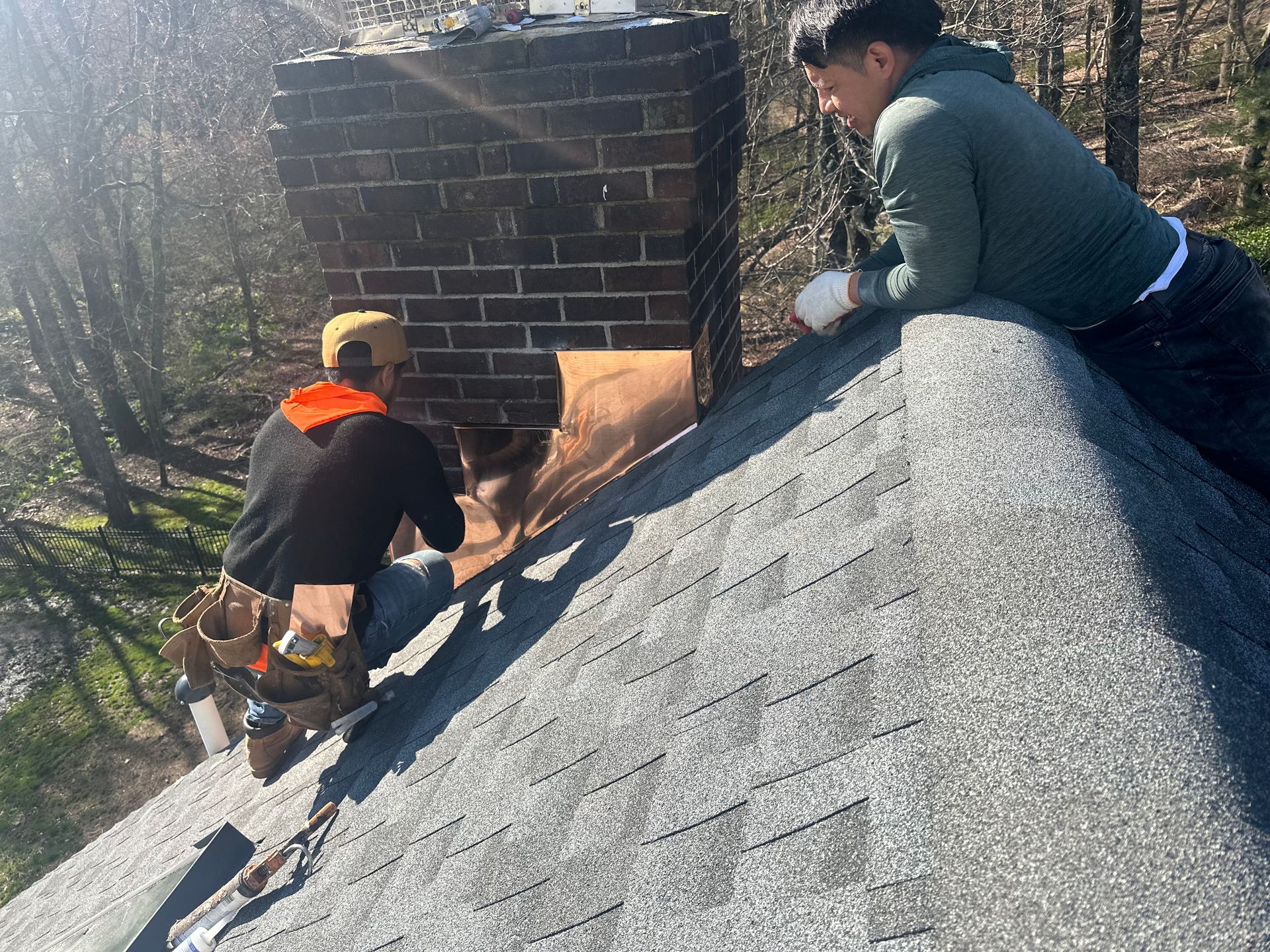 Two people work on a brick chimney on a shingled roof, one reaching into an opening while the other observes.