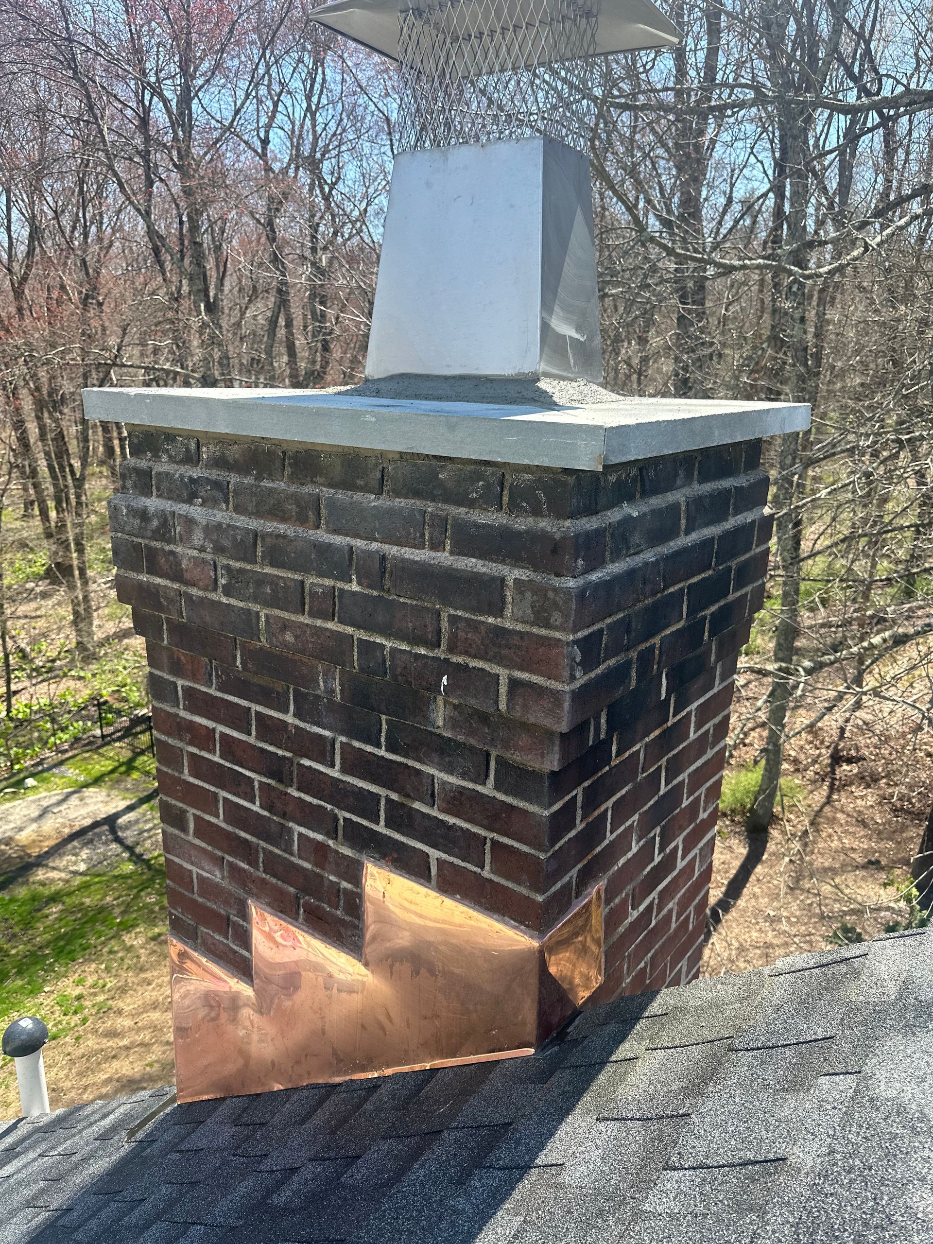 A brick chimney on a shingled roof with a copper flashing base and a metal cap against a background of trees.