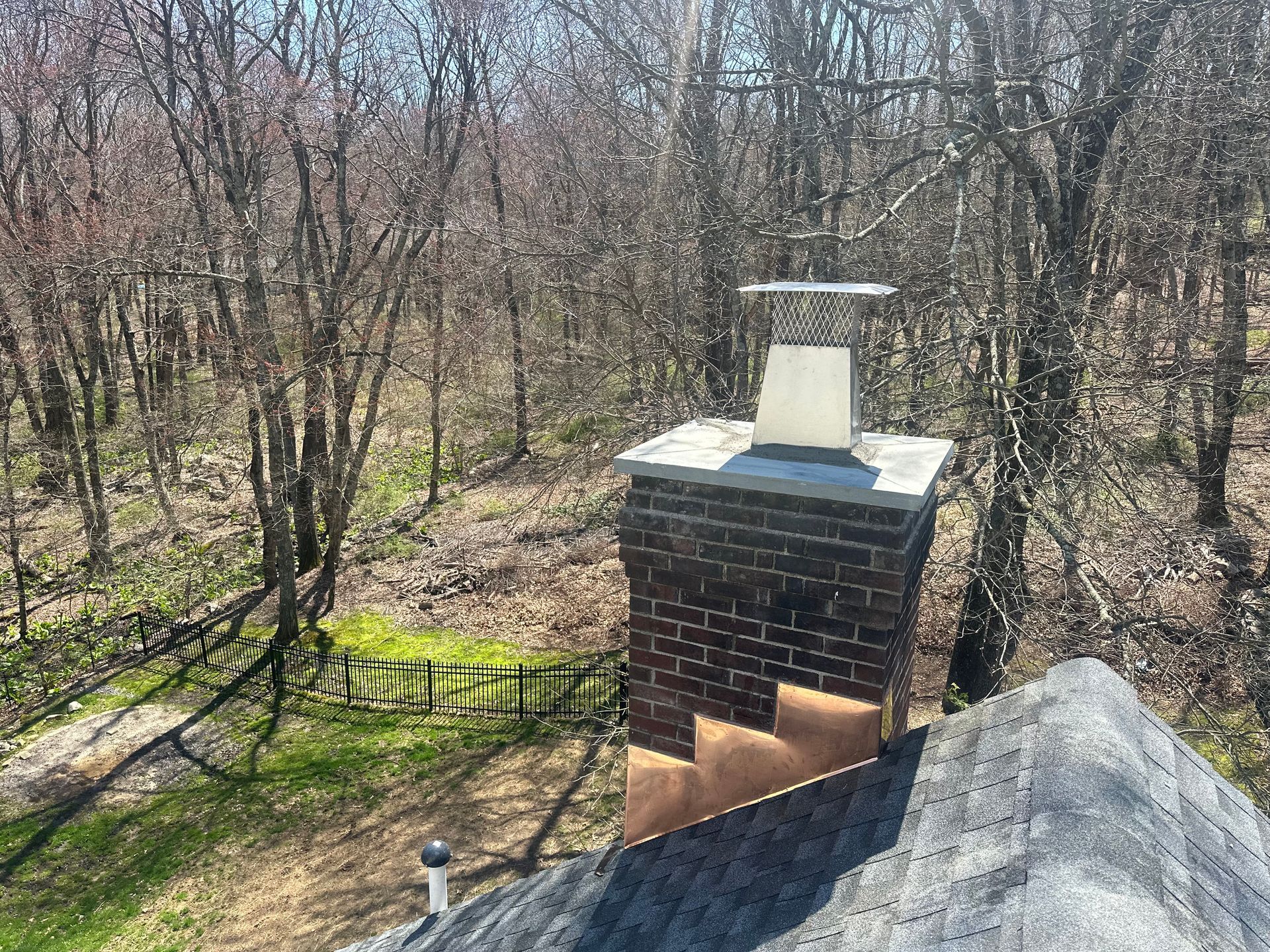 A brick chimney with a metal cap and copper flashing stands on a shingled roof against a backdrop of bare trees.