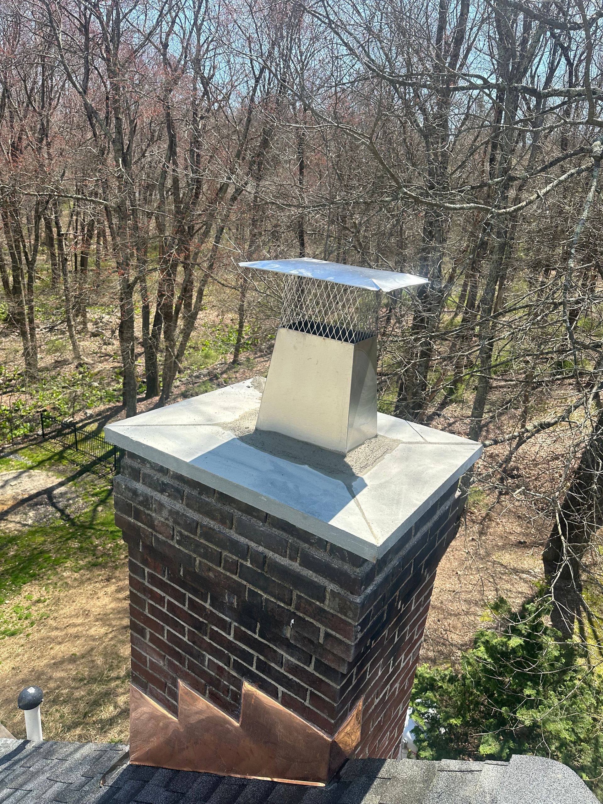A stainless steel chimney cap and vent centered on a brick chimney with copper flashing, set against a backdrop of trees.