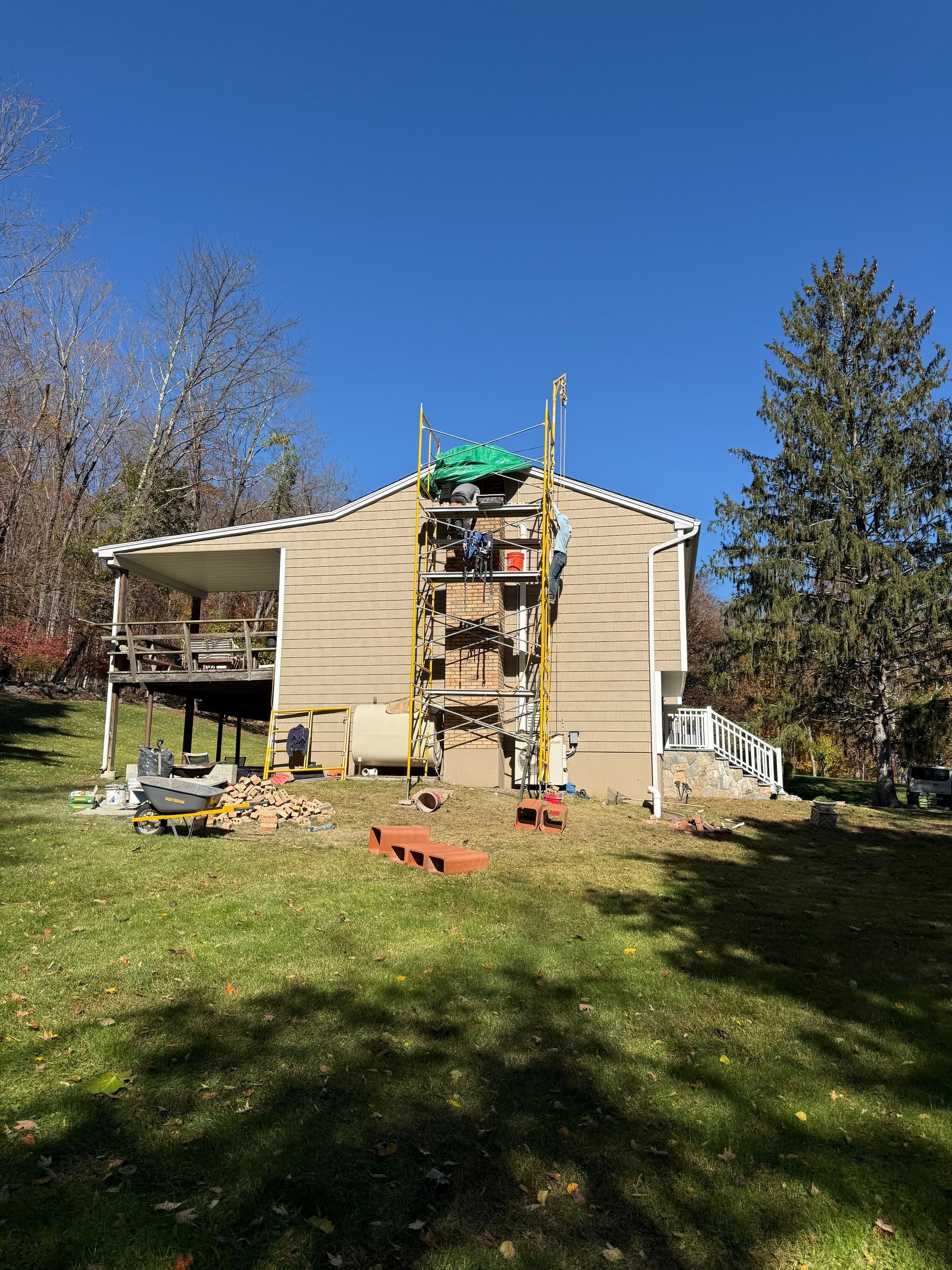 A tan house side featuring scaffolding and a partially dismantled chimney on a sunny day with trees in the background.