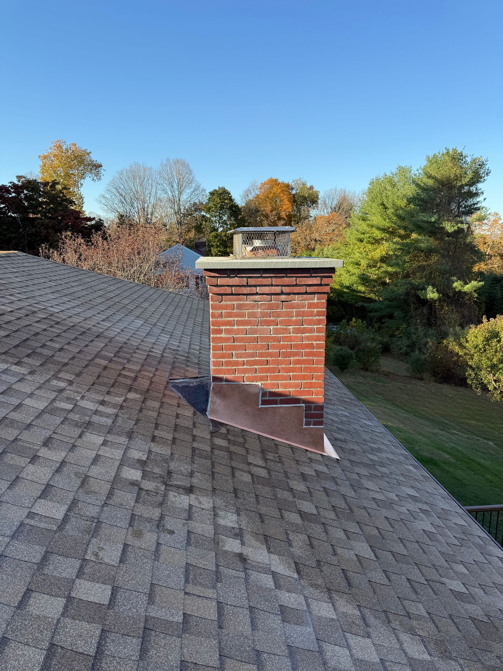 A brick chimney with copper flashing stands on a shingled roof under a clear blue sky with autumn trees in the background.