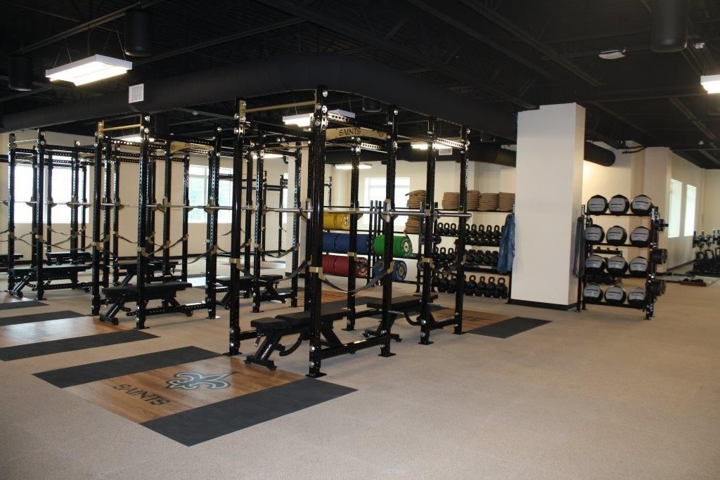 Weightlifting gym with weight racks, benches, and free weights. Beige floor, black ceiling, white pillars.