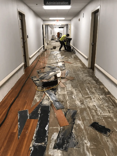 Hallway floor being renovated; worker operating floor grinder; damaged floorboards and debris.
