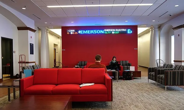 A red sofa faces a red wall with a digital sign. Two people sit at a table. Light-colored walls and carpet.