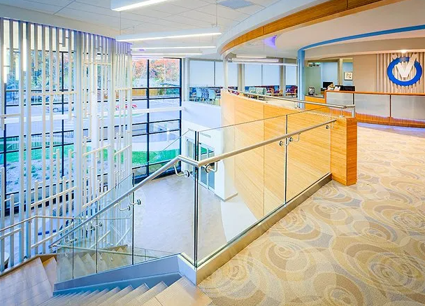 Staircase with glass railing, leading to a light-filled lobby with reception desk and decorative wall.