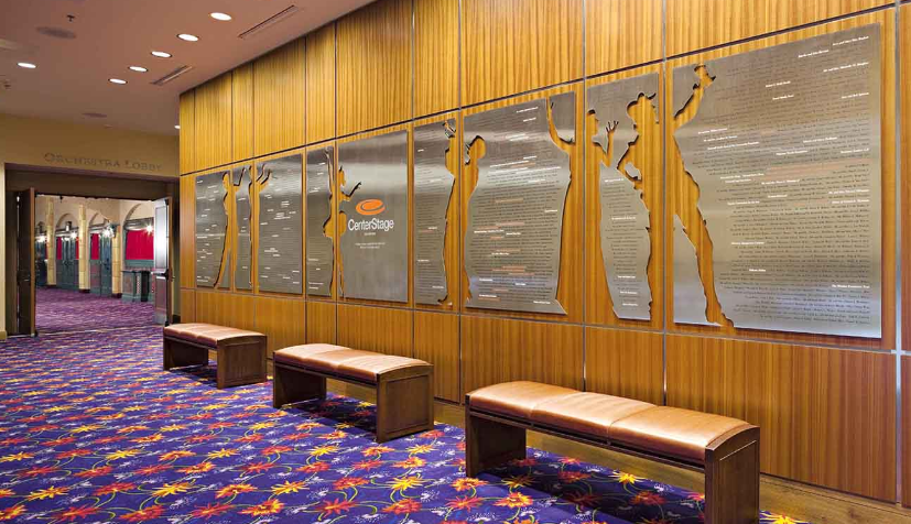 Hallway with wooden walls, benches, and a carpet with a colorful floral pattern. Decorative panels with figures are on the wall.