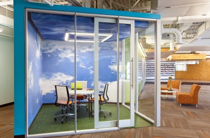 Modern office meeting room with sky-themed walls, glass doors, a round table, and orange chairs.