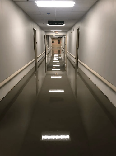 Long hallway with reflective, dark floor. White walls, doors, and lights. Handrails along the walls.