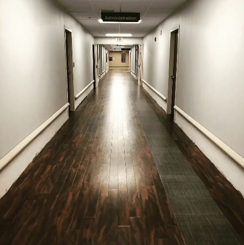 Long, empty hospital hallway with wood floor, doors, and a sign above a door that reads 