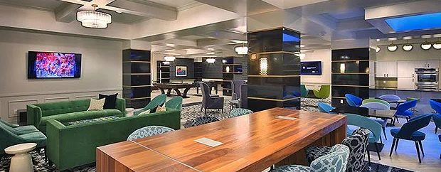 A lounge area with green couches, blue chairs, a large wooden table, and a TV on the wall.