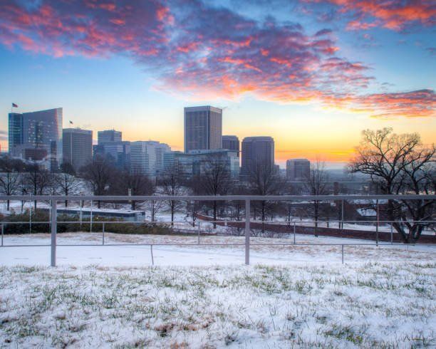 Snowy landscape with a city skyline at sunrise. Pink, orange, and blue sky; white snow; bare trees.